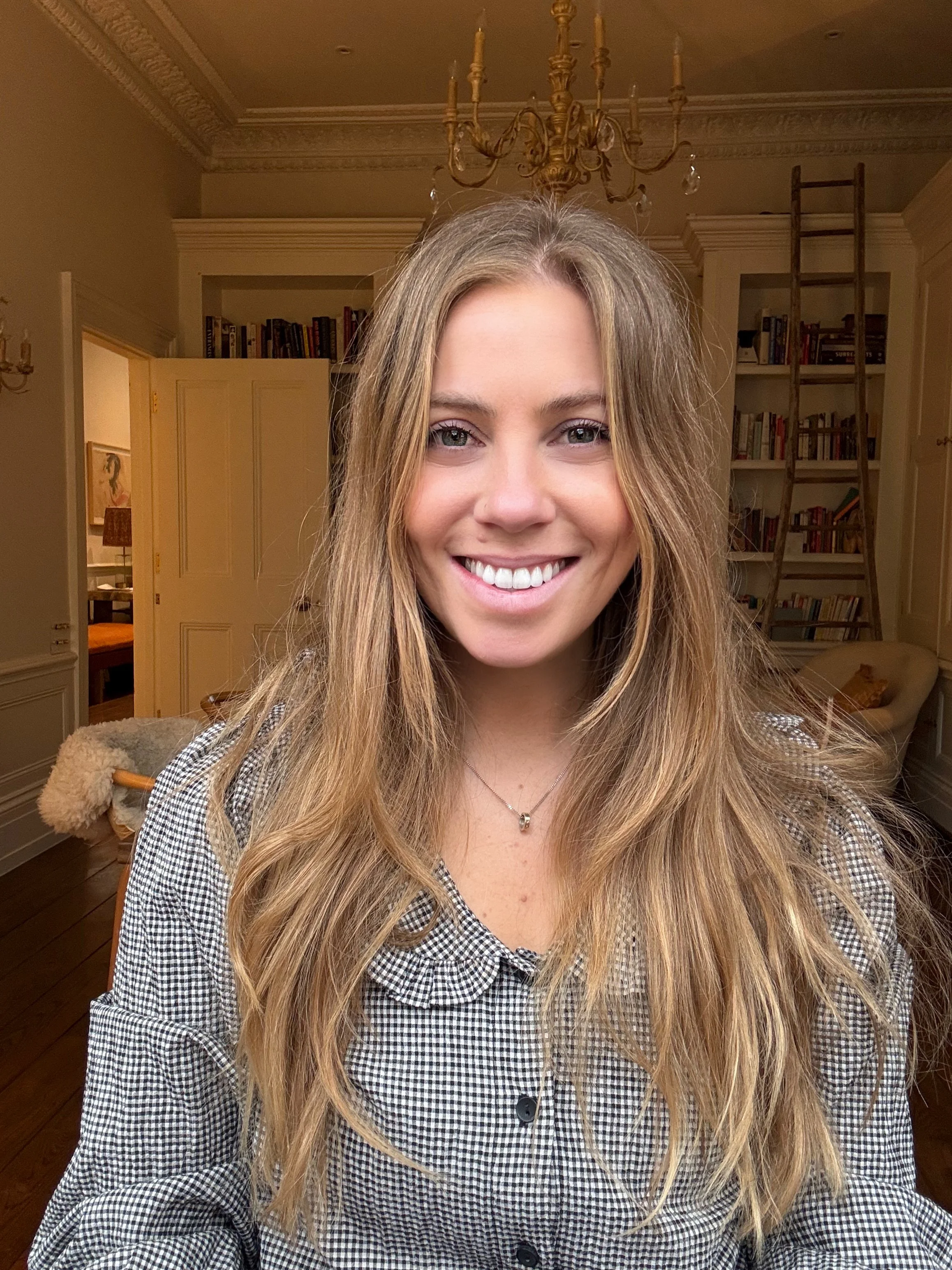 A young woman with long, wavy blonde hair smiling at the camera in a cozy, well-lit living room with bookshelves, a ladder, and a chandelier overhead.