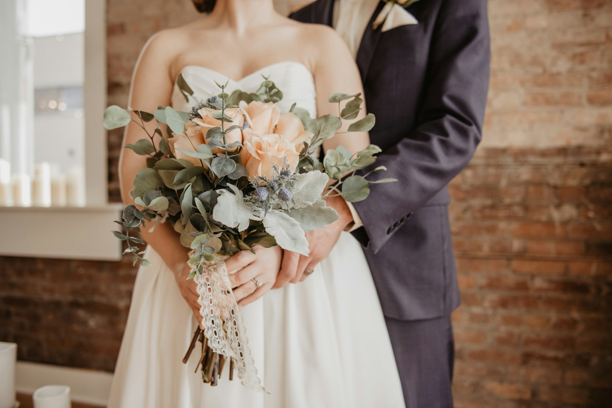 Une mariée tenant un bouquet de fleurs roses et vertes, avec un homme en costume sombre en arrière-plan, dans une salle avec mur en briques et bougies sur le rebord de la fenêtre.