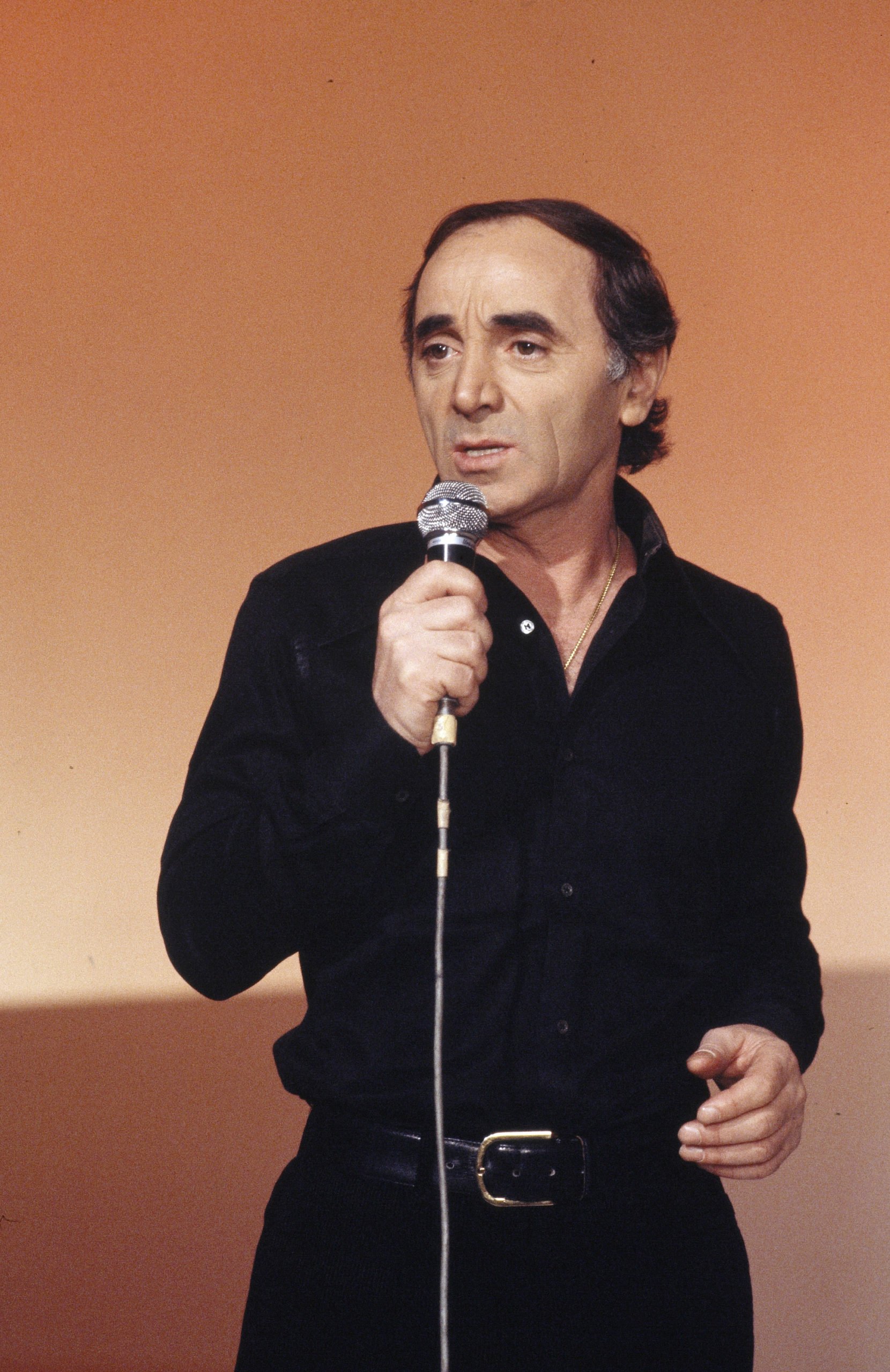 Un homme avec des cheveux foncés, portant une chemise noire, tenant un microphone, faisant une performance ou un discours, devant un fond orange.