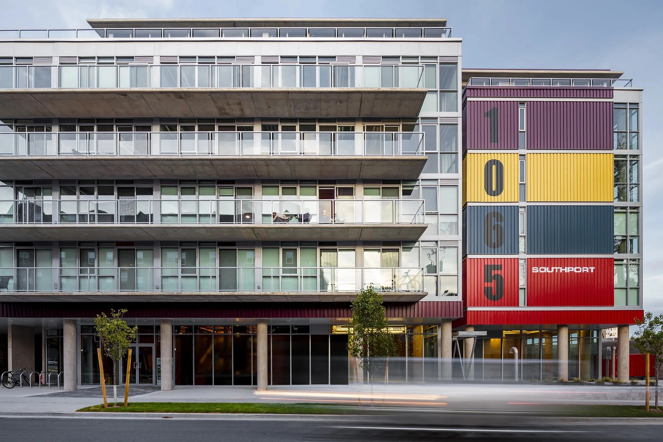Modern multi-story residential building with colorful shipping container-style accents on the right side labeled 1 to 6, and the word SOUTHPORT on one red container. The building features glass balconies, concrete pillars, and trees in the foreground, with light streaks indicating moving traffic.