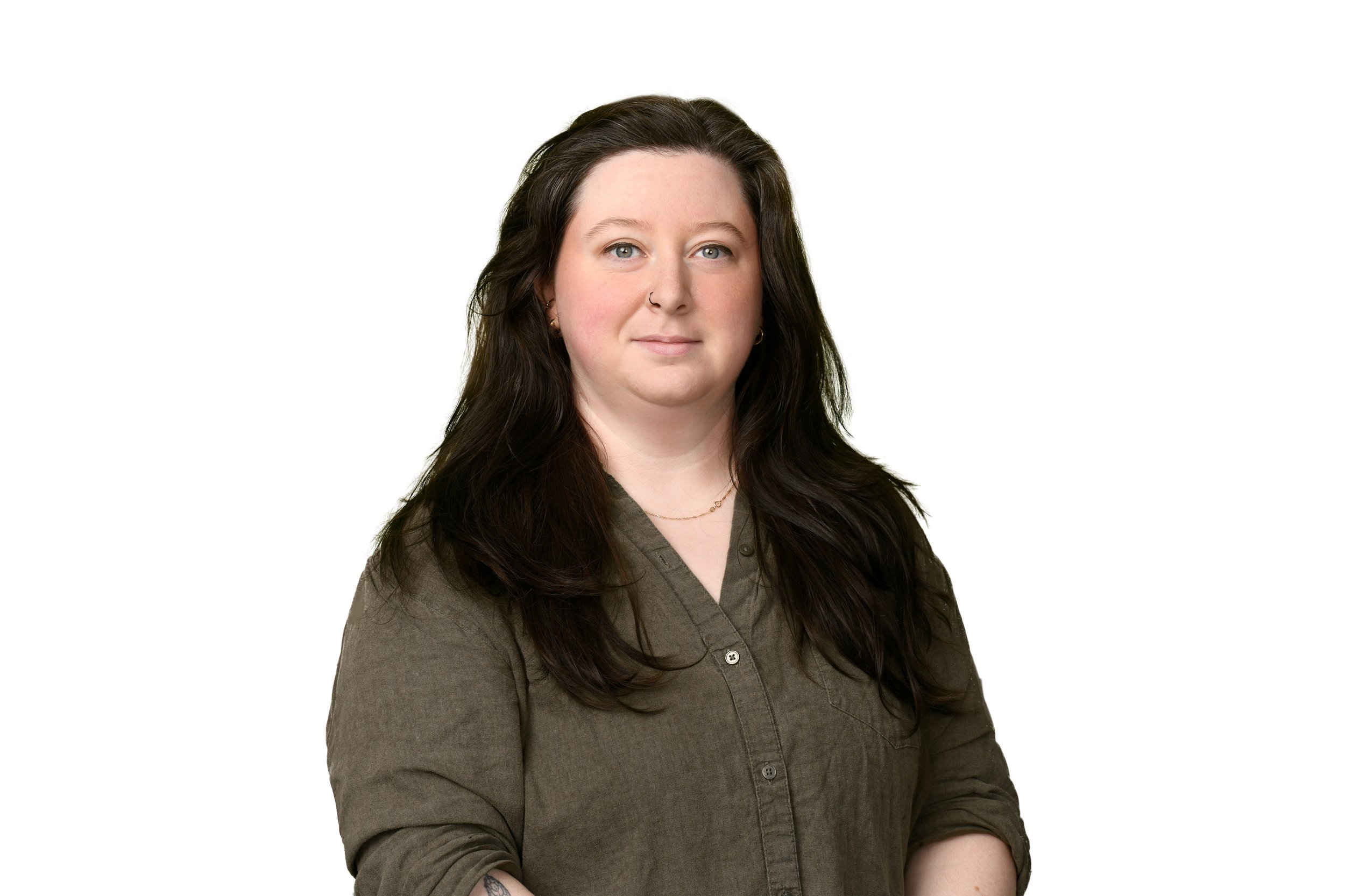 A woman with long dark hair, blue eyes, wearing a dark olive green shirt and a delicate gold necklace, stands against a plain white background.