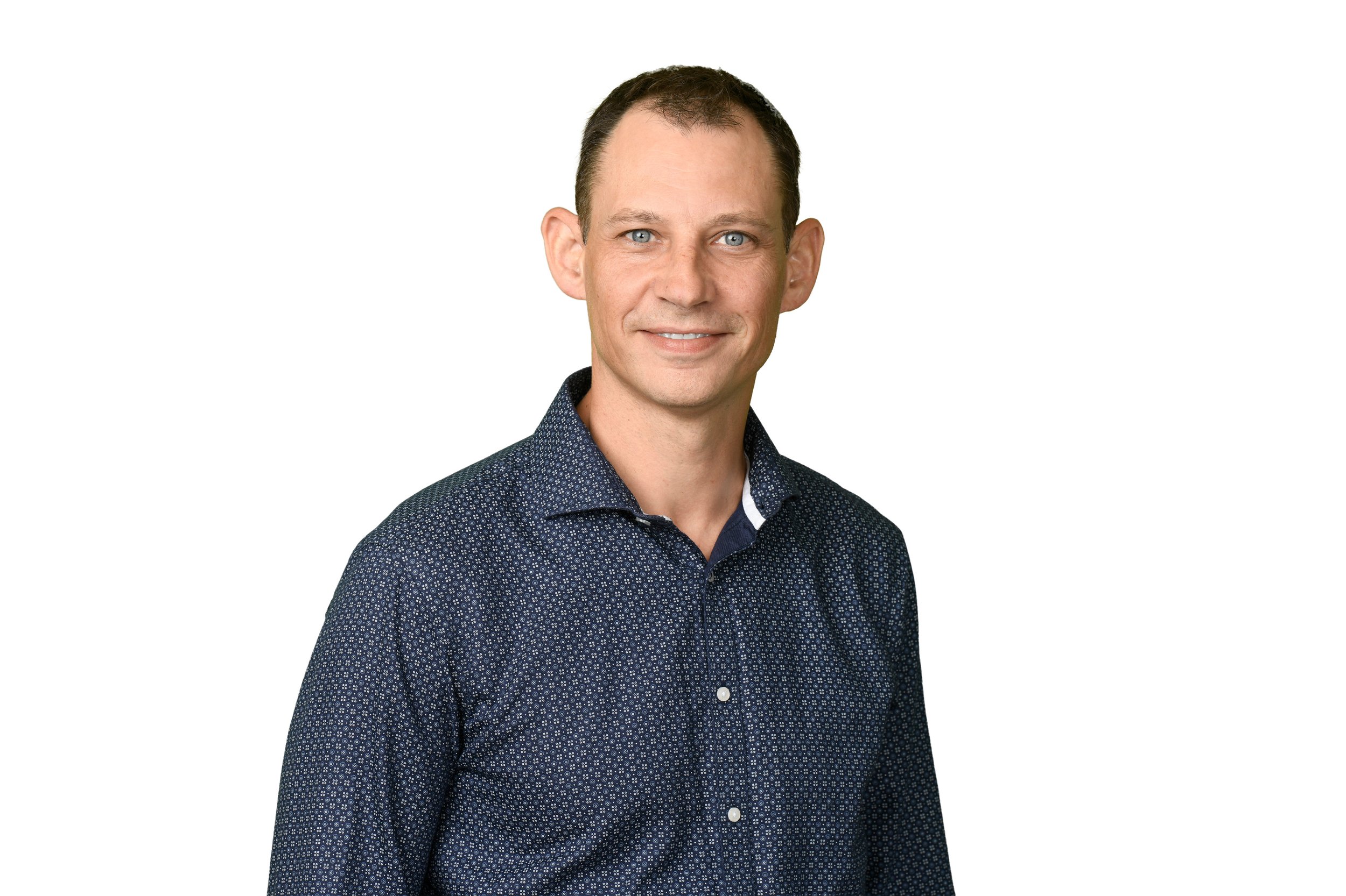 Head and shoulders portrait of a smiling man with short dark hair, wearing a navy blue patterned button-up shirt, against a plain white background.