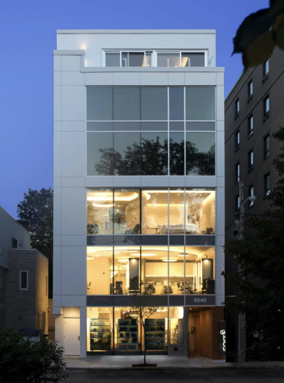 Modern multi-story office building with large glass windows, illuminated interior, and street view in the evening.