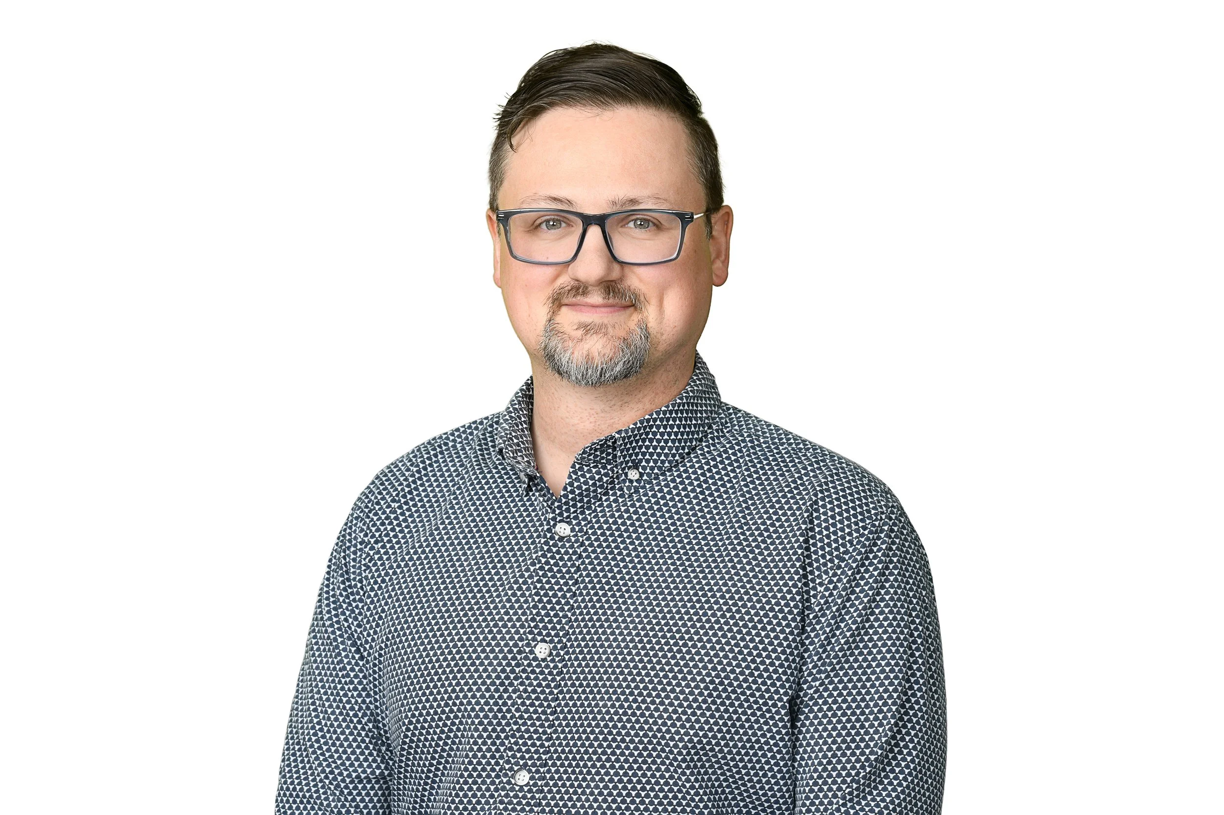 A man with glasses, short brown hair, and a beard, wearing a patterned button-up shirt, posing against a plain white background.