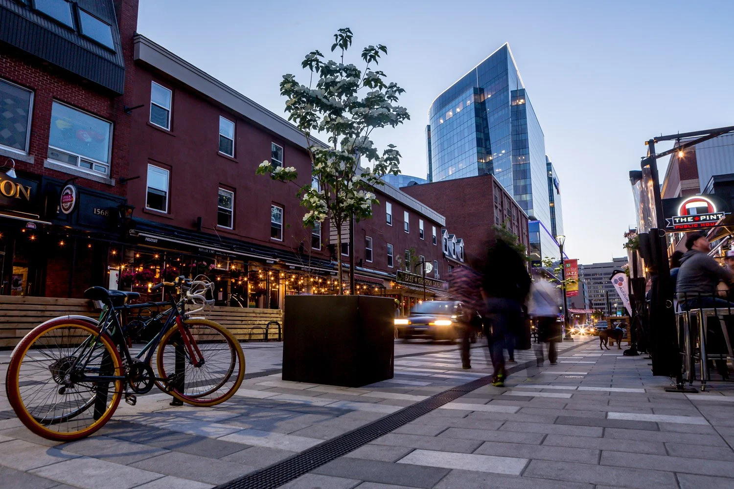 City street during dusk with people walking, a bicycle parked on the sidewalk, a building with lit-up string lights, and modern glass skyscrapers in the background.