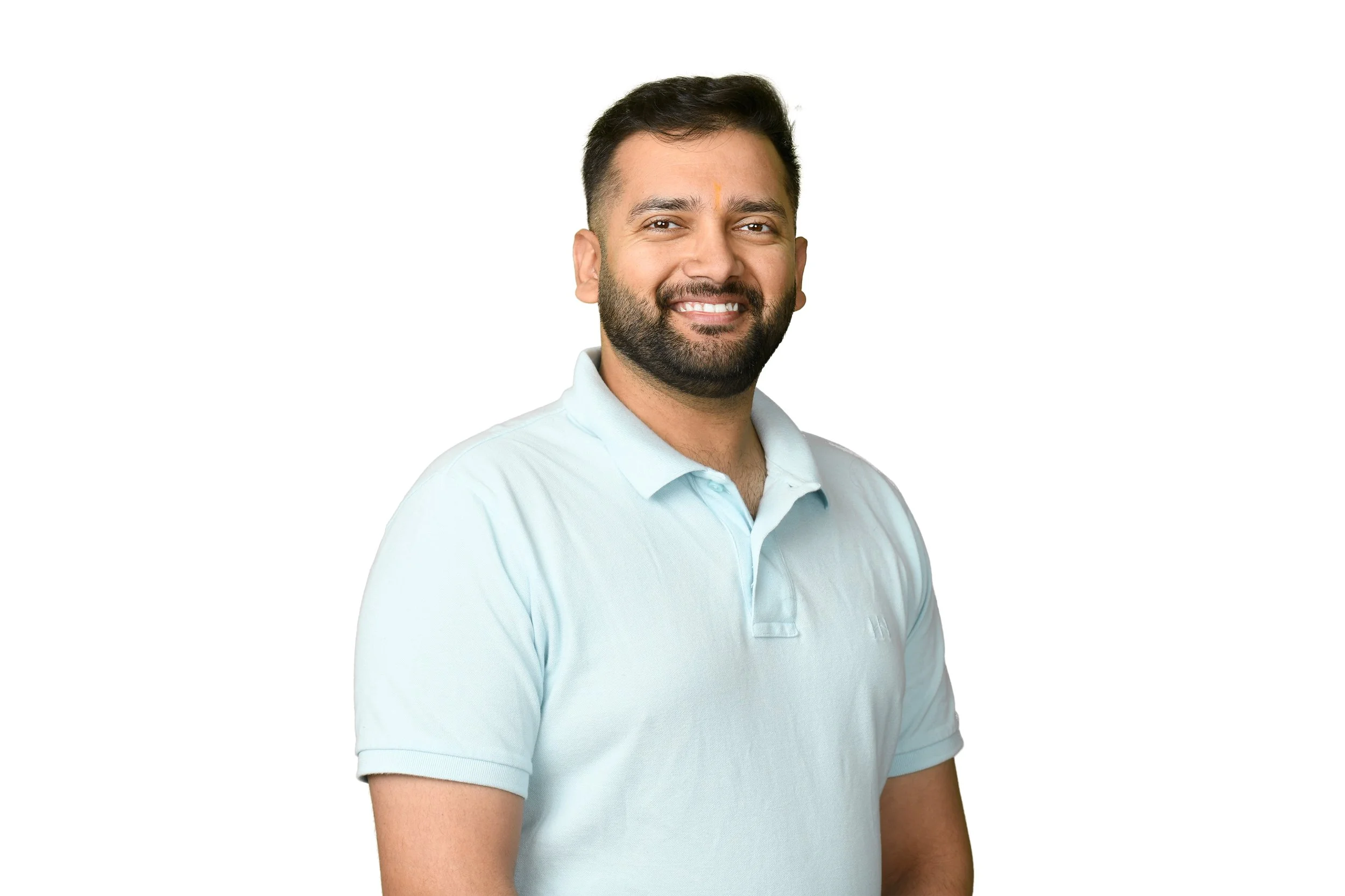 A smiling man with a beard and short dark hair, wearing a light blue polo shirt, standing against a plain white background.