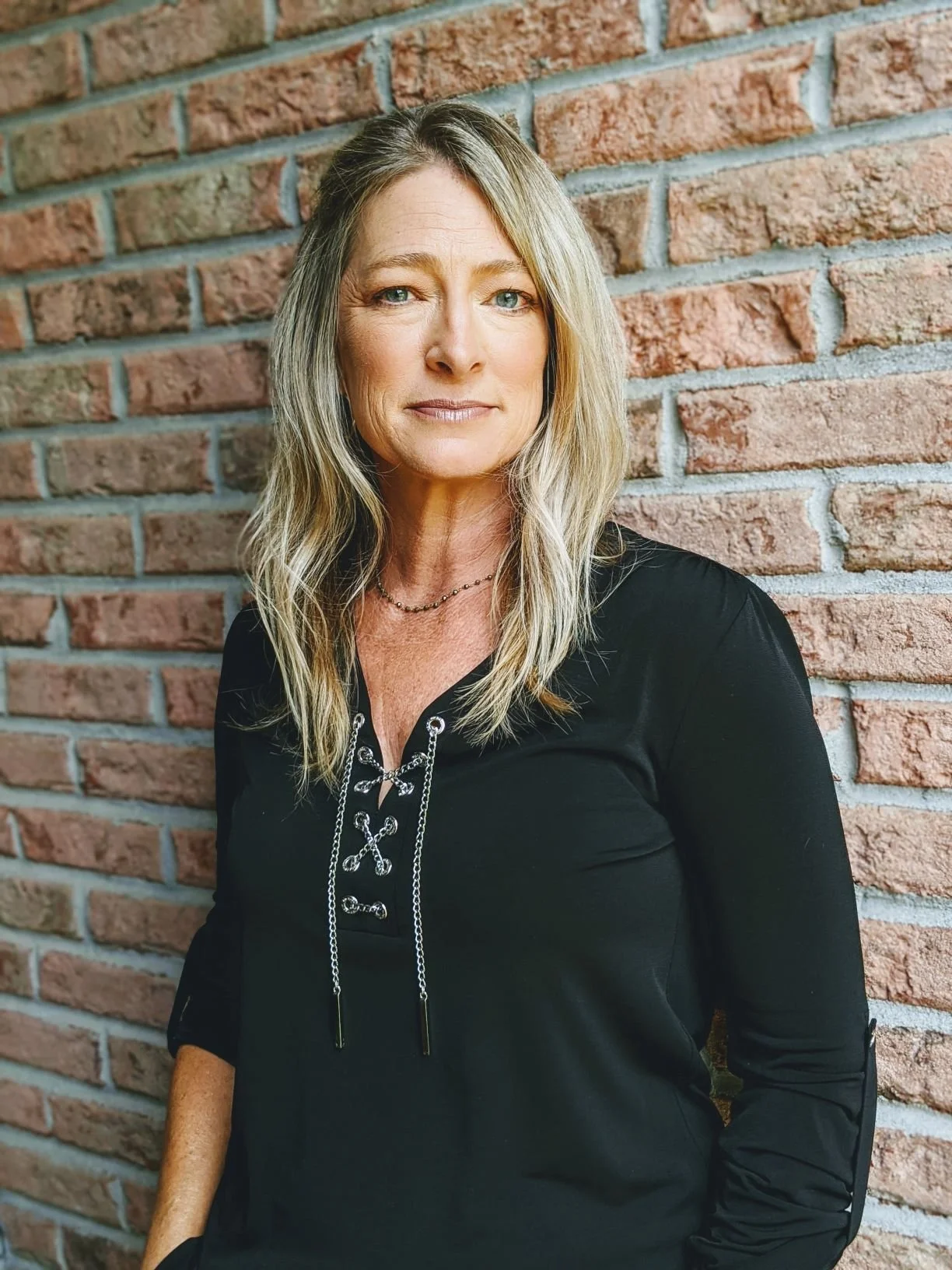 A middle-aged woman with blonde hair and blue eyes, wearing a black top with lace-up detail, posing against a brick wall.