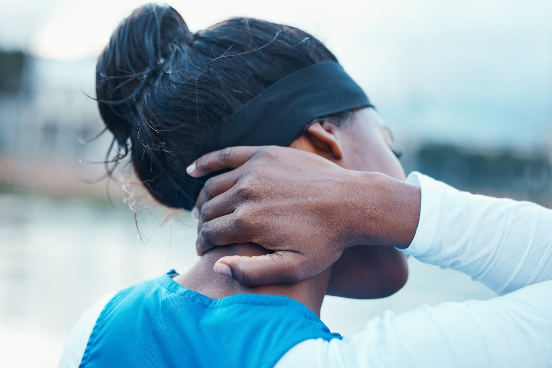 A person with dark hair in a bun wearing a black headband and a white and blue athletic shirt holds their neck in discomfort near a body of water.