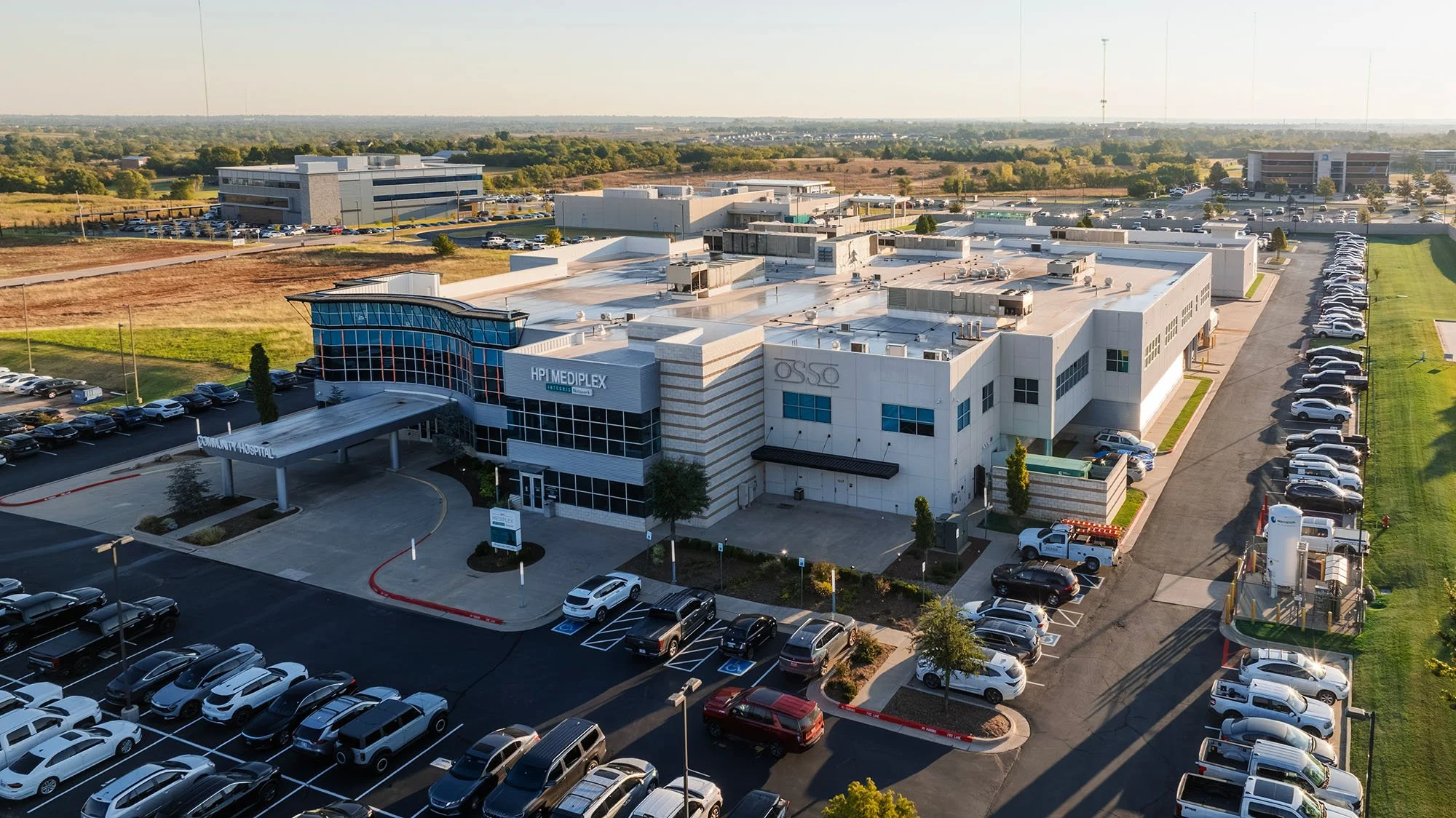 Aerial view of a hospital complex with parking lots, surrounded by open fields and a few office buildings in the background.