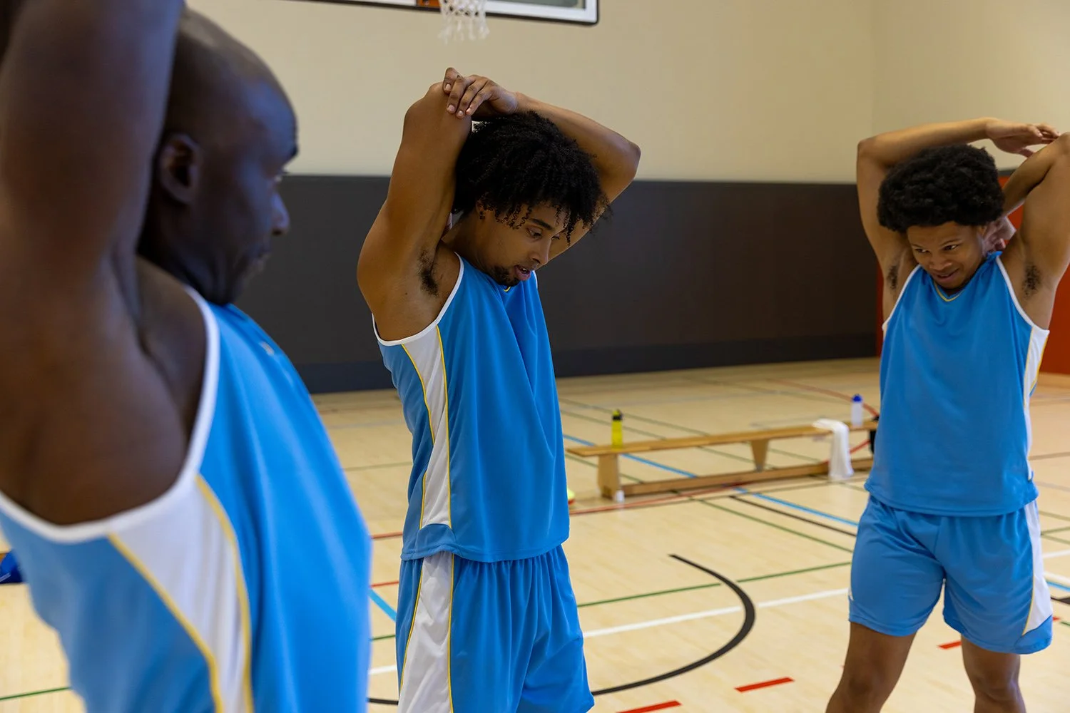 Four male basketball players in blue uniforms stretching on an indoor basketball court.
