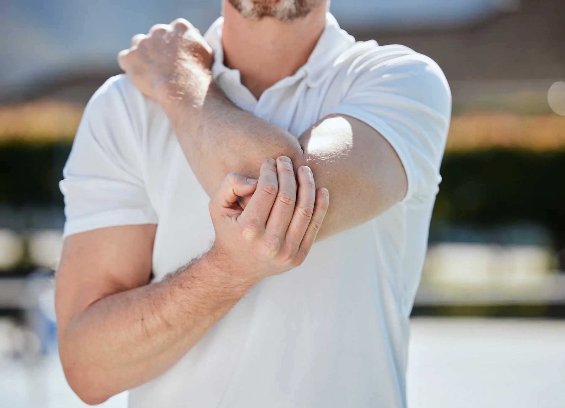 A man in a white polo shirt is stretching or massaging his arm outdoors on a sunny day.
