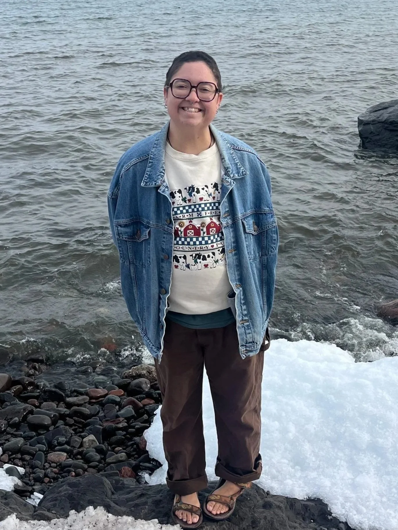 Person smiling and standing on a rocky beach with water behind them, wearing a denim jacket, black shorts, and sandals.