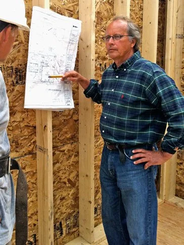 A construction supervisor in a plaid shirt and glasses discusses building plans on a construction site, pointing at blueprints attached to a wooden stud wall.