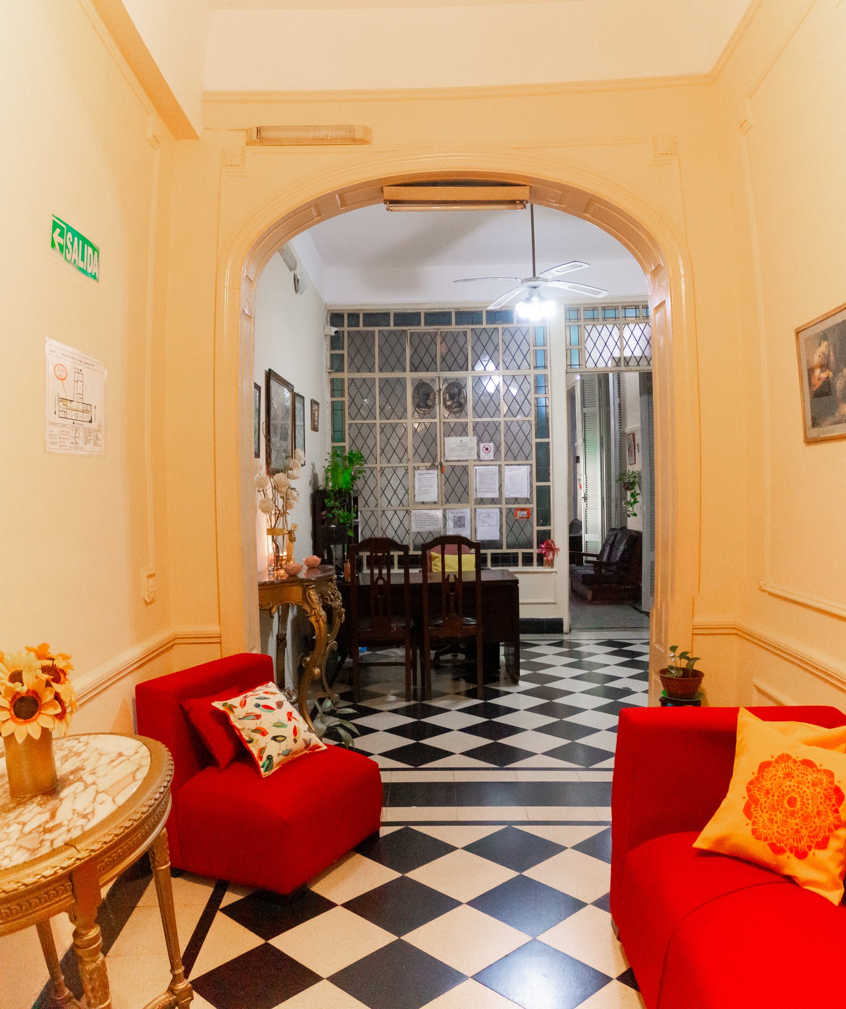 Sala con paredes amarillas, muebles de madera oscura, sillones rojos con cojines decorativos, una mesa de mármol, plantas, cuadros en las paredes, ventanales con rejas y un ventilador en el techo.