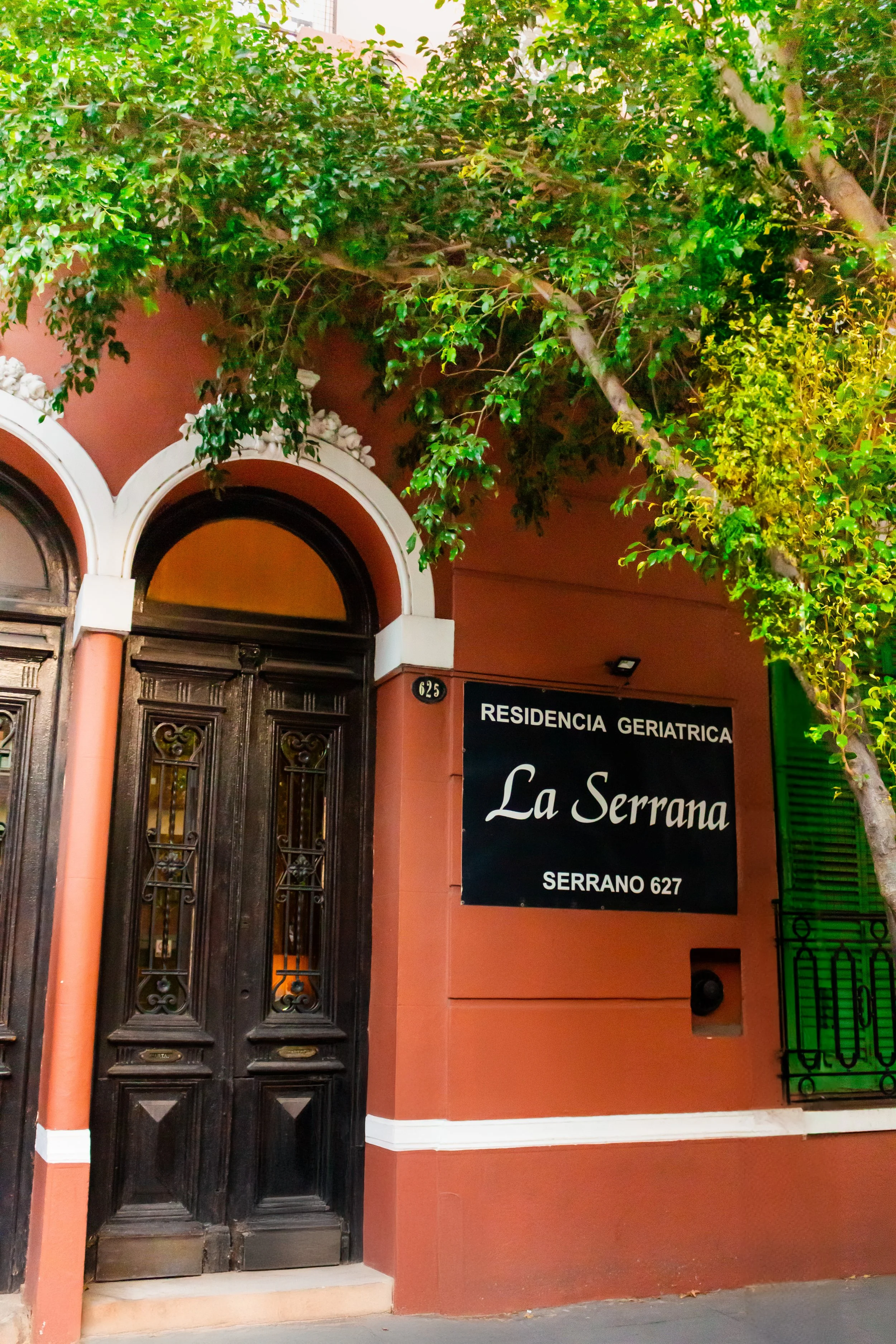 Entrada de una residencia geriátrica llamada La Serrana, ubicada en Serrano 627, con puerta de madera oscura y un árbol verde en la fachada.