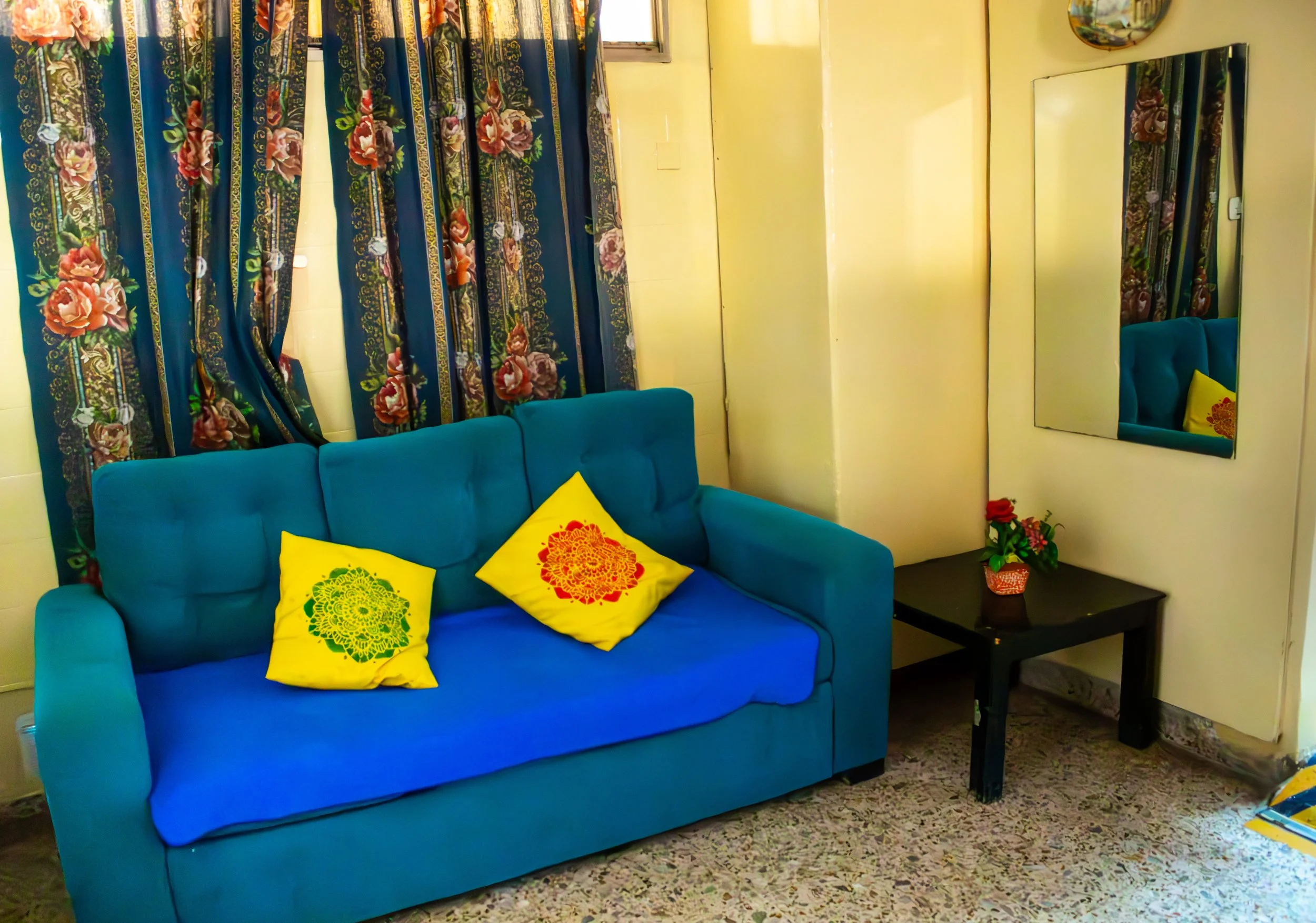 Sala con sofá azul, cojines amarillos con diseño de flor, cortinas con flores y patrones en tonos rosas, verdes y dorados, espejo en la pared, ventana y mesa de centro con planta decorativa.