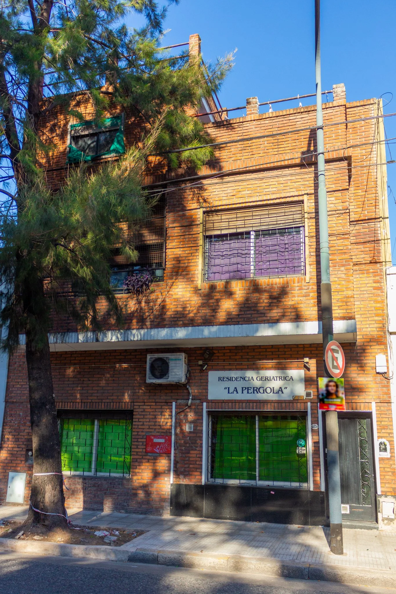 Edificio de ladrillos con cartel que dice 'Residencia Geriátrica La Pergola', ventanas con cortinas moradas y verdes, y un árbol en primer plano.