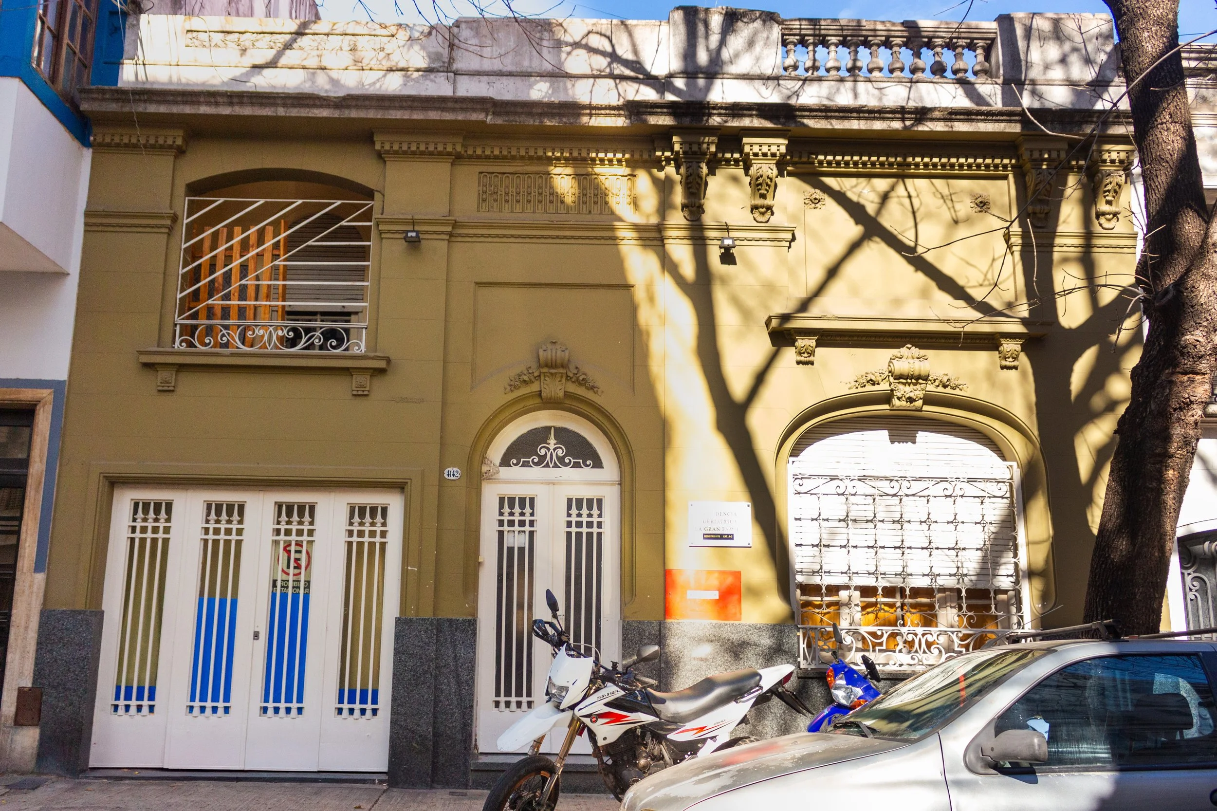 fachada de edificio residencial con puertas y ventanas de rejas, árboles en frente, motocicletas y coche estacionados en la calle