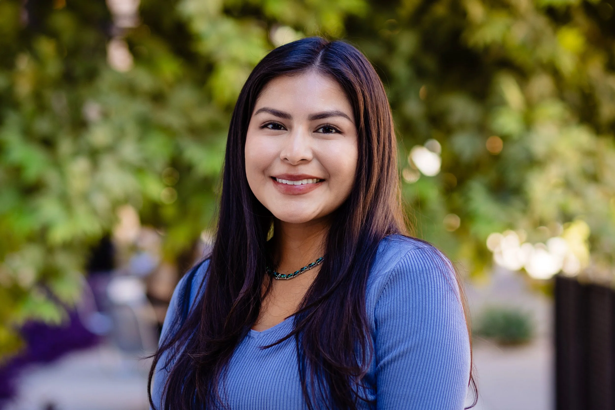 Caleigh smiling in a blue blouse with green foliage in the background