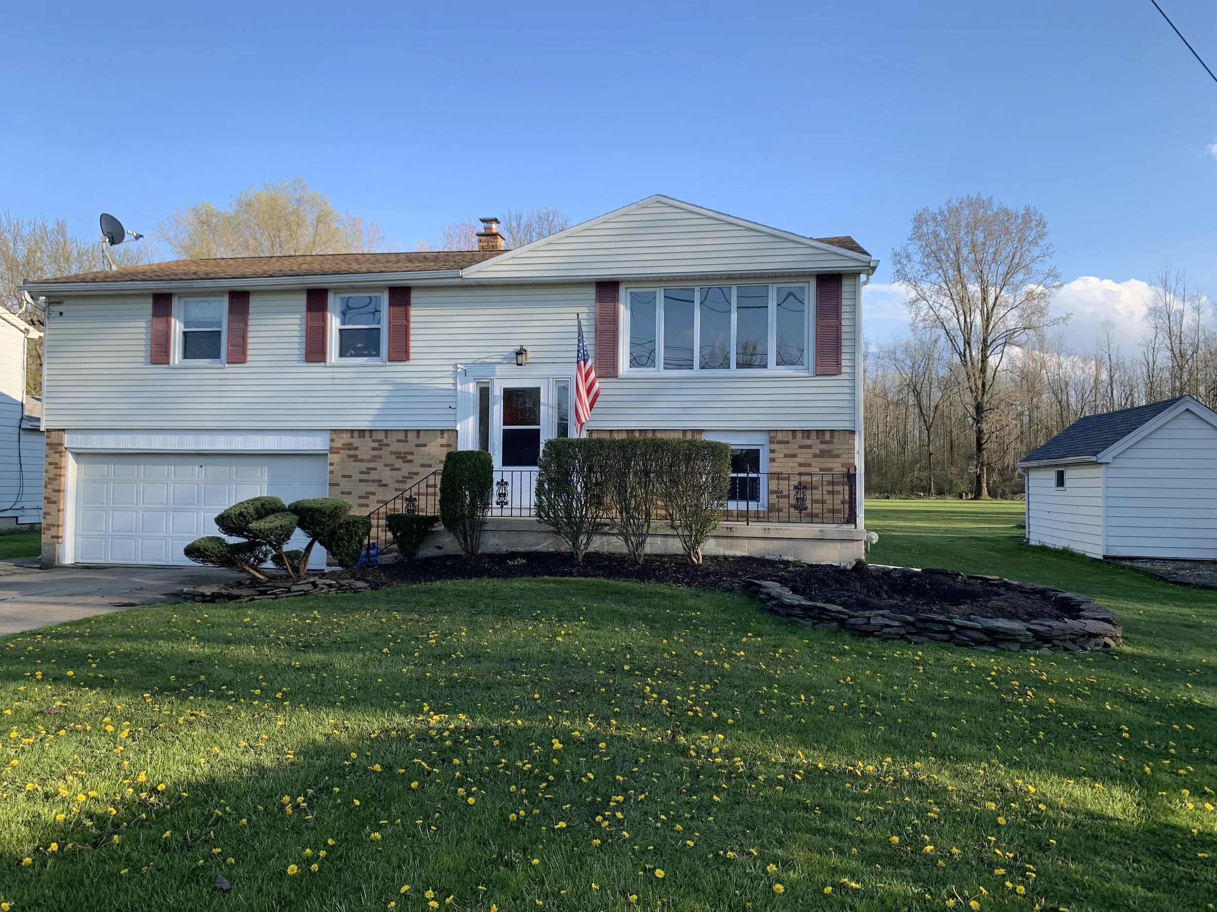 A two-story house with a brick and white siding exterior, an attached garage, landscaped front yard with trimmed bushes, and an American flag by the front door. The yard has green grass with small yellow flowers and a stone-lined garden bed. There ar
