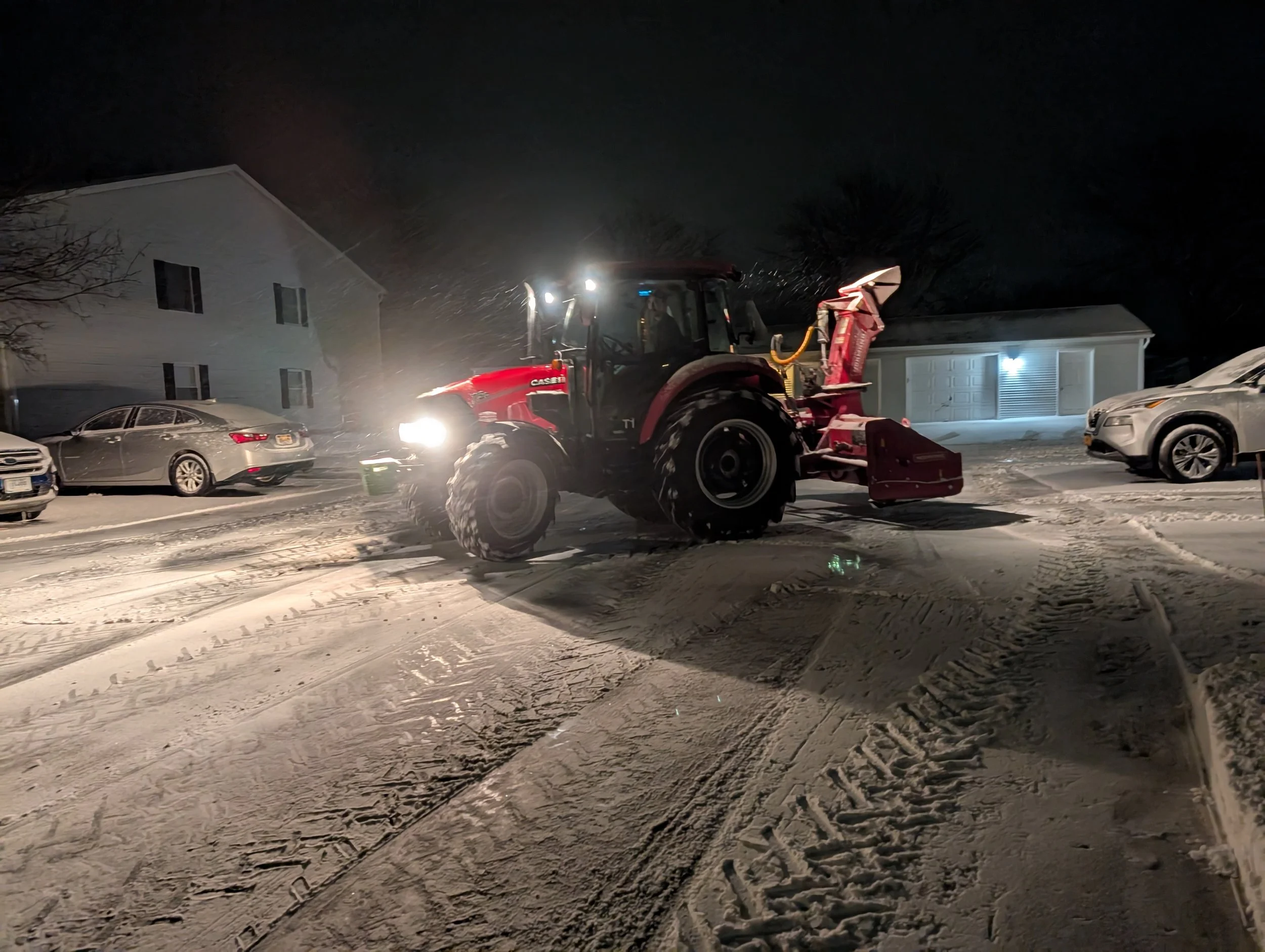 A red snow plow clearing snow from a residential street at night, with snow-covered parked cars and houses in the background.