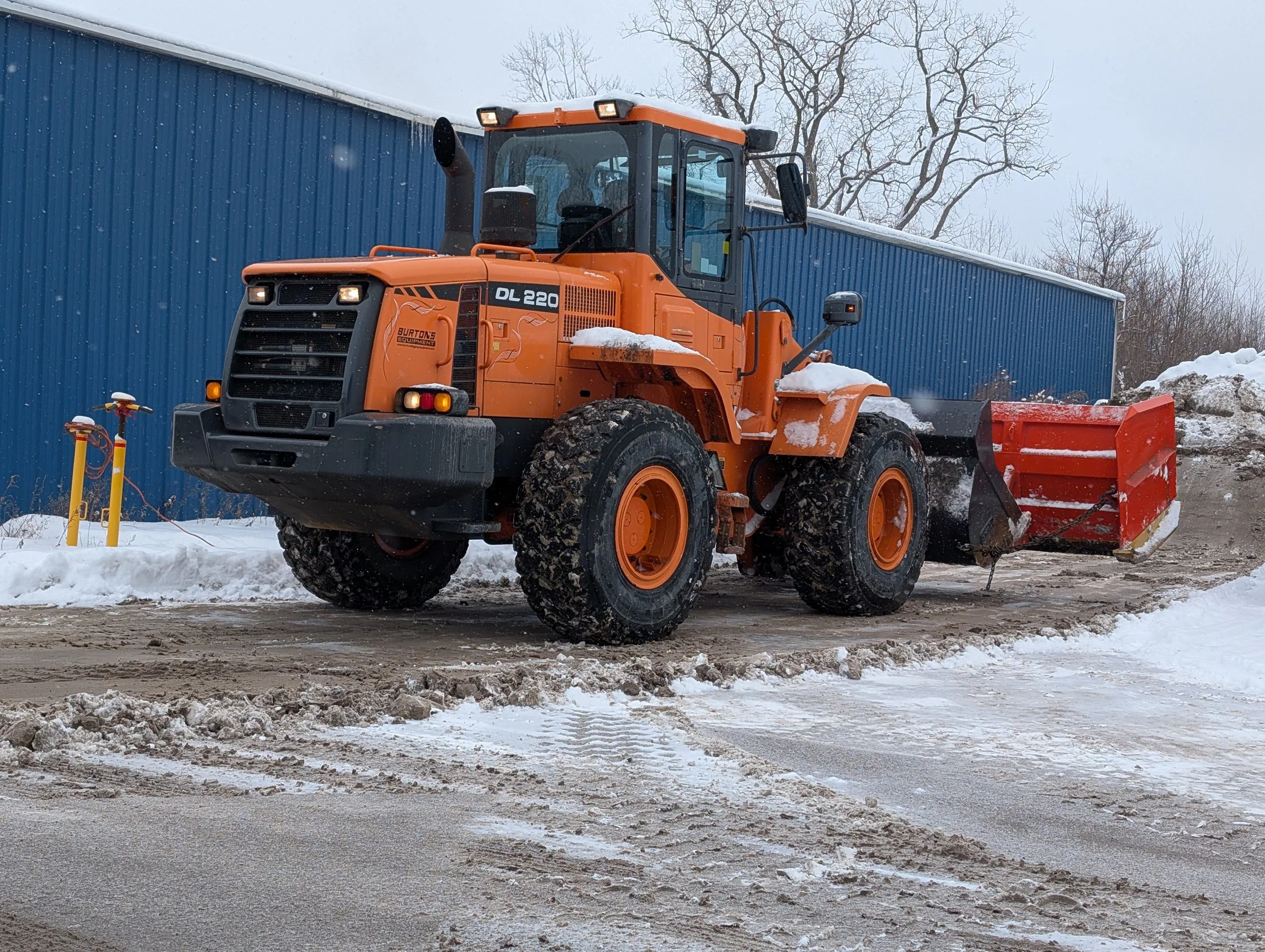 Orange snowplow clearing snow on a dirt-covered road with a blue building and snow piles in the background.