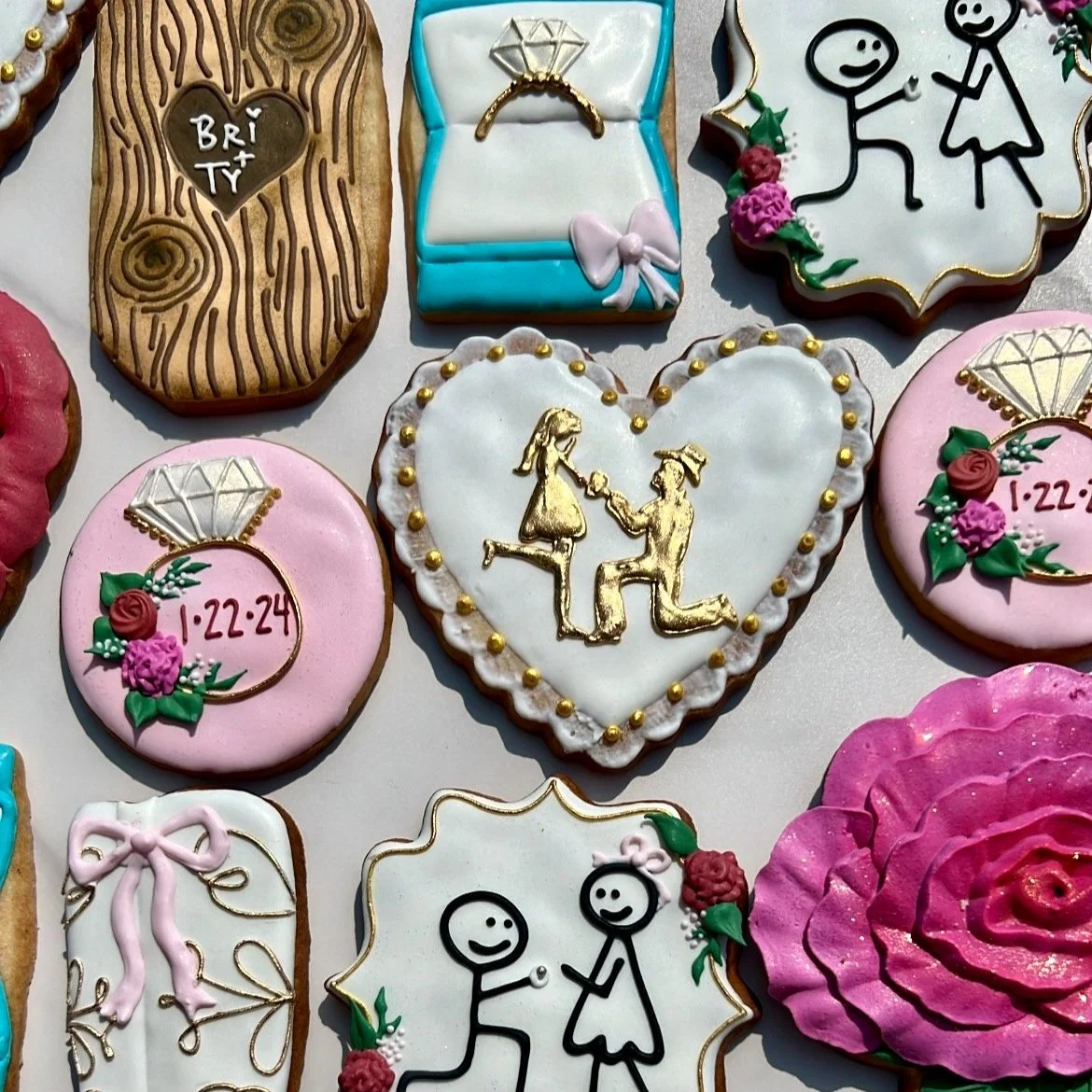 Assorted decorated cookies, including wedding, engagement, and love-themed designs with hearts, rings, stick-figure couples, and date markings.