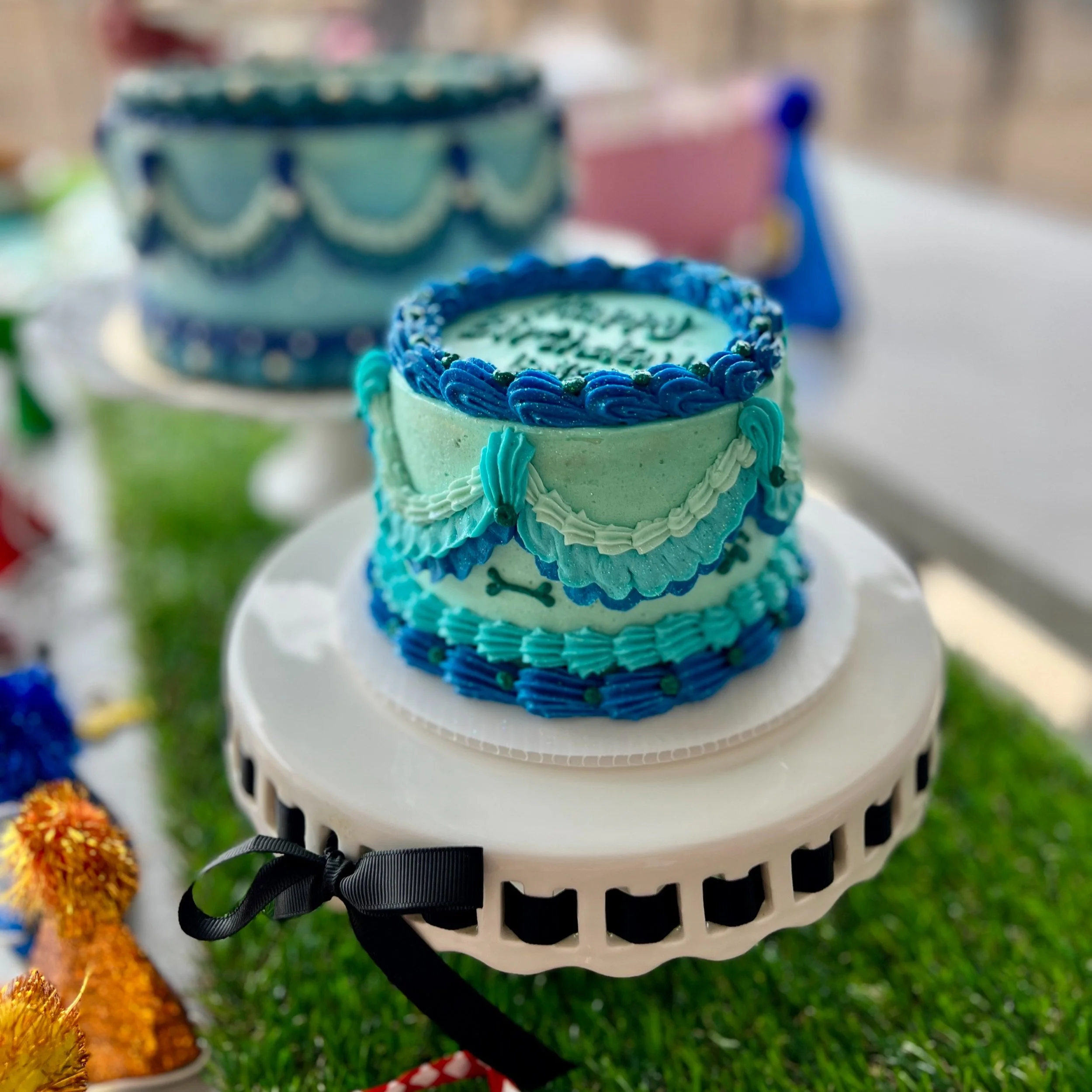 A two-tiered blue birthday cake decorated with piping and a dog bone design, placed on a white cake stand with a black ribbon, with colorful decorations and other cakes in the background.