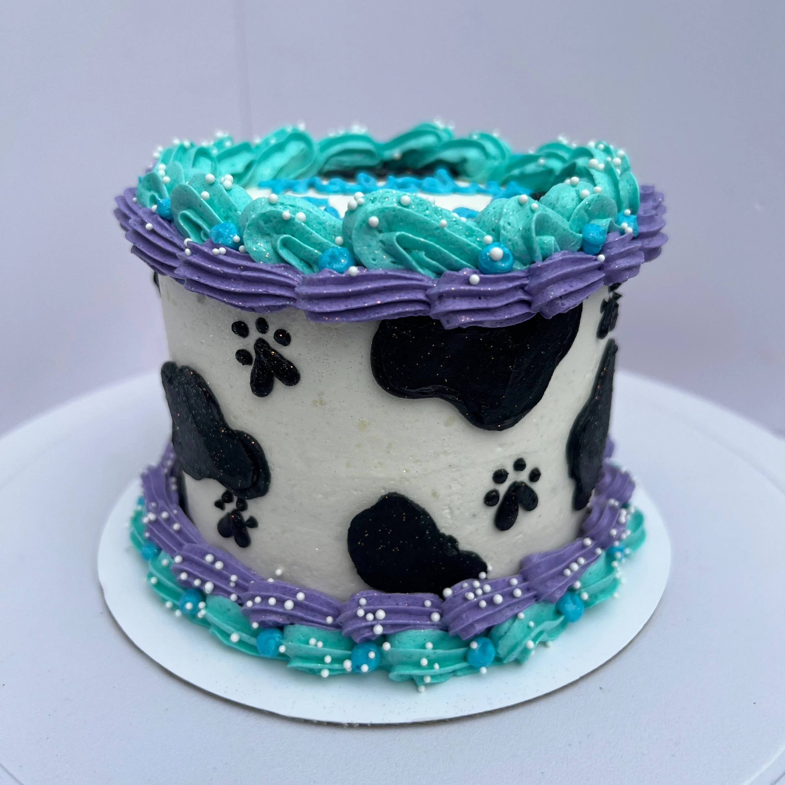 Cake decorated with black and white cow print pattern, purple and turquoise frosting borders, and turquoise frosting on top with white and turquoise pearls.
