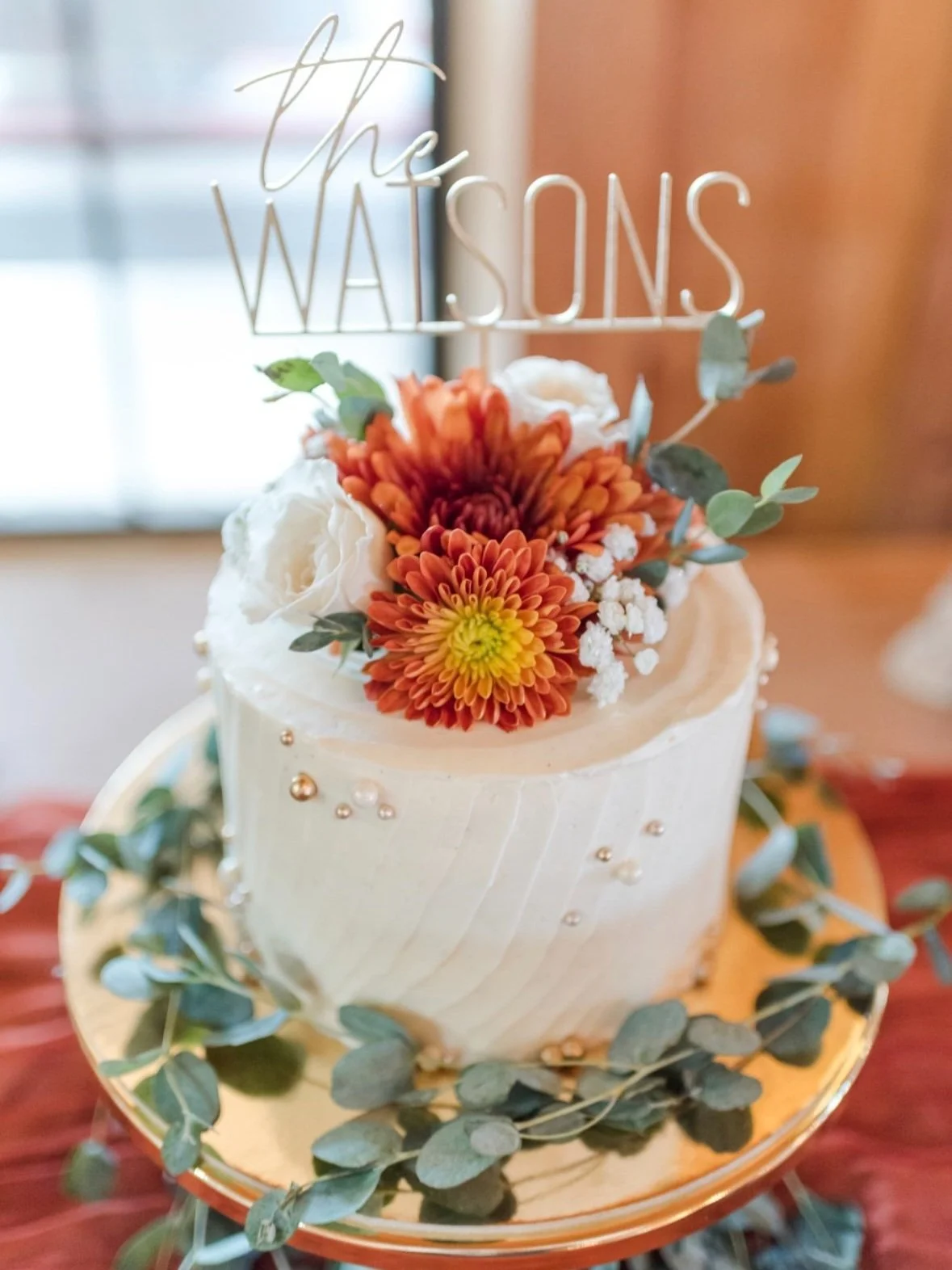 Wedding cake decorated with white frosting, orange and yellow flowers, greenery, and a topper that reads "The Watsons."