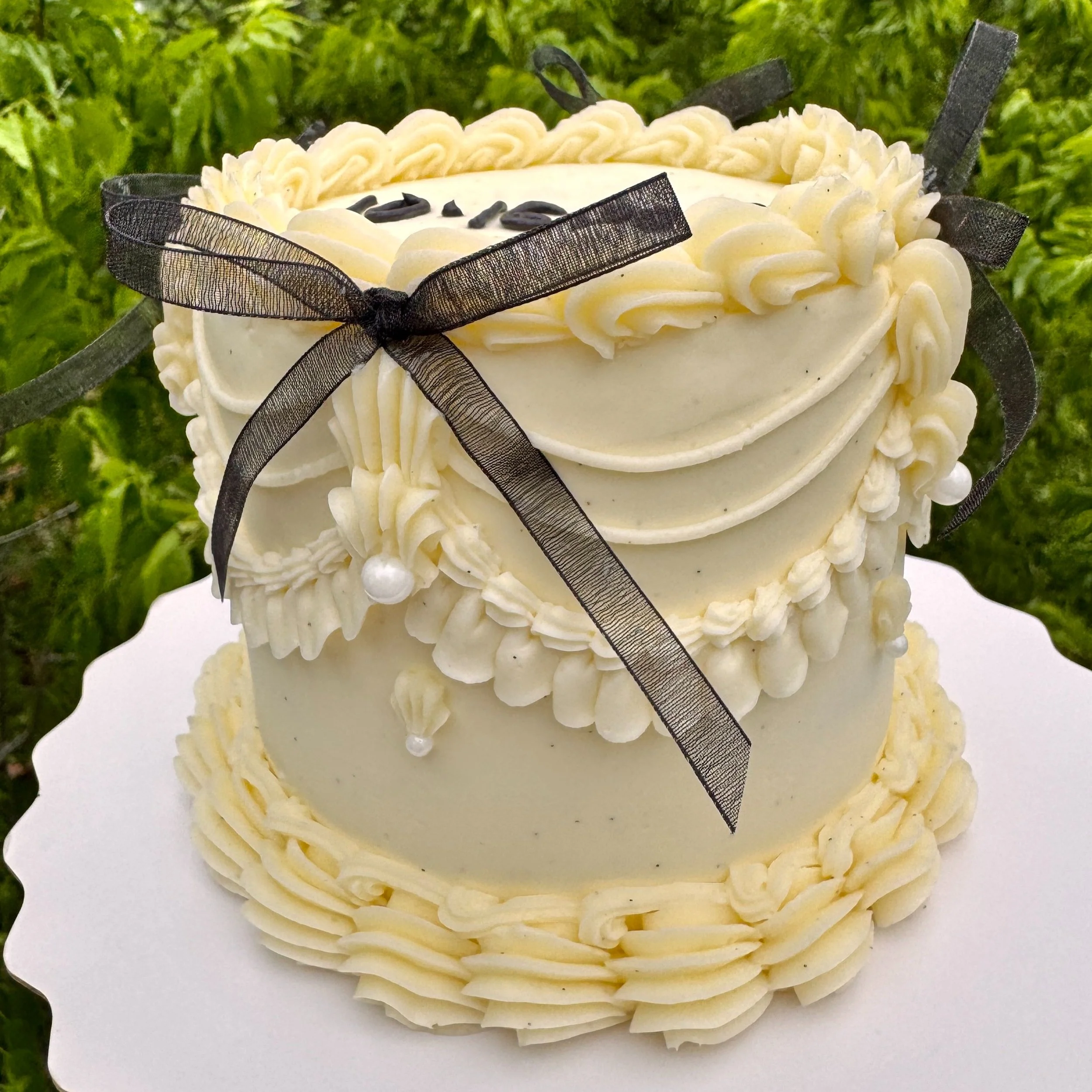 A heart-shaped white cake decorated with cream piping, pearls, and a black ribbon bow, set against a green leafy background.