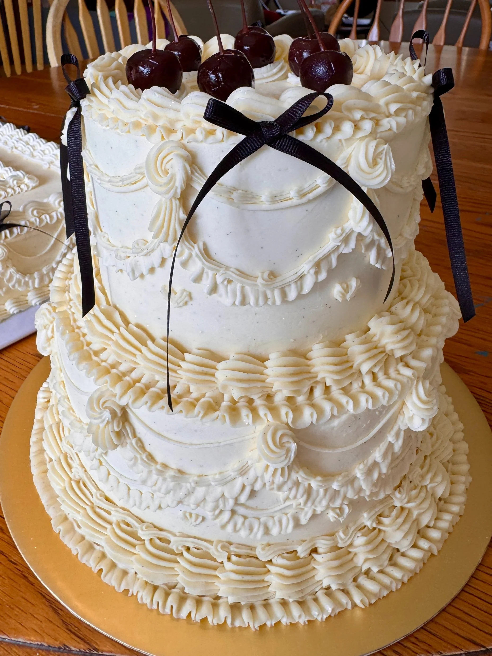 Three-tier white wedding cake decorated with piped white icing and topped with red cherries, black ribbons, and decorative icing patterns.