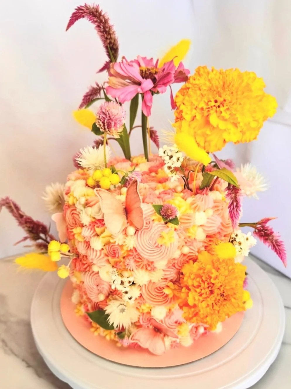A floral cake decorated with pink, yellow, and white flowers and a butterfly, featuring pink frosting in rosette patterns, on a white cake stand.