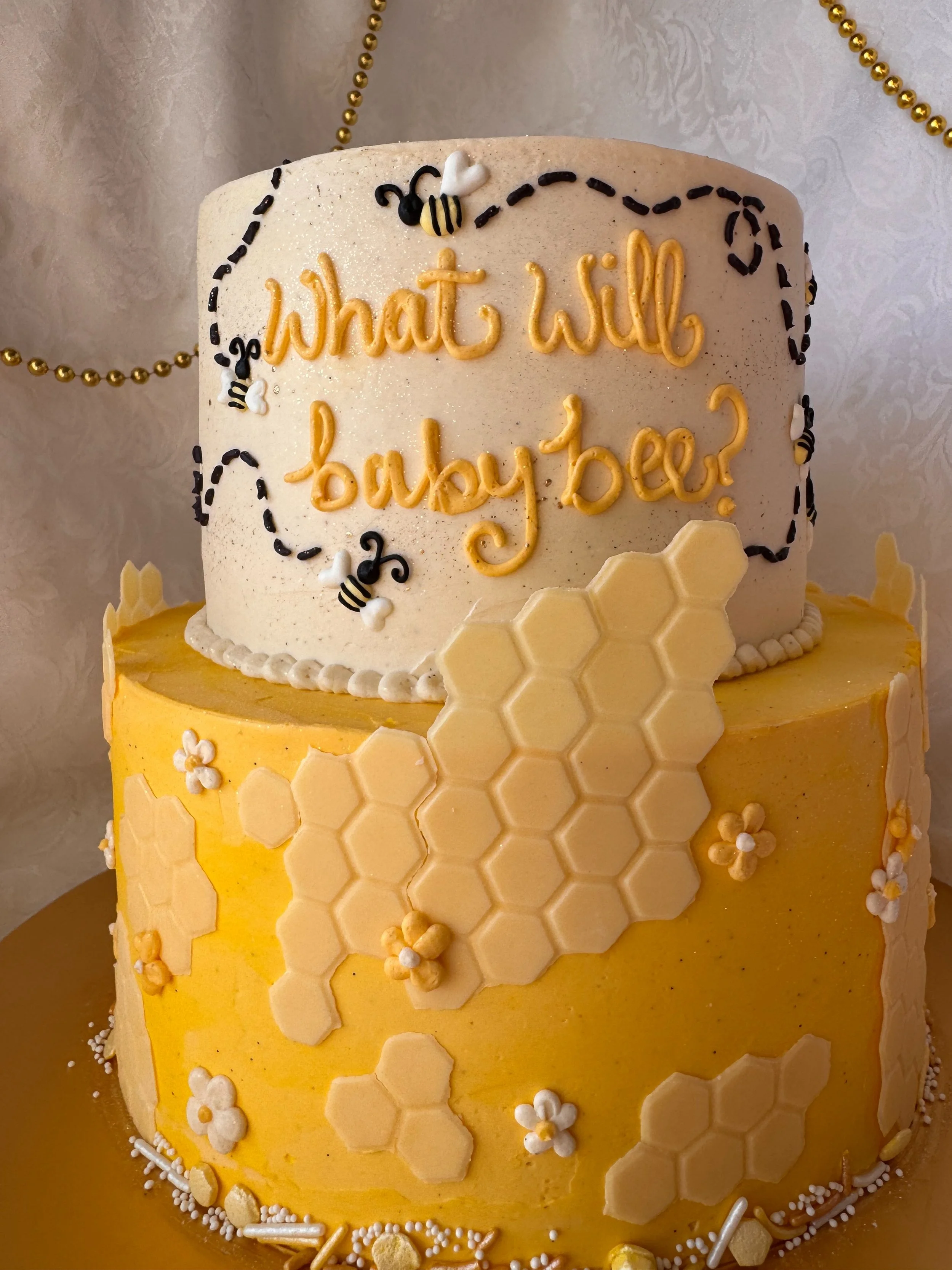 A two-tiered cake with the top tier white and decorated with small bee and heart-shaped decorations, and the bottom tier yellow decorated with honeycomb and flower designs. The top tier has the words 'What will baby bee?' written on it.