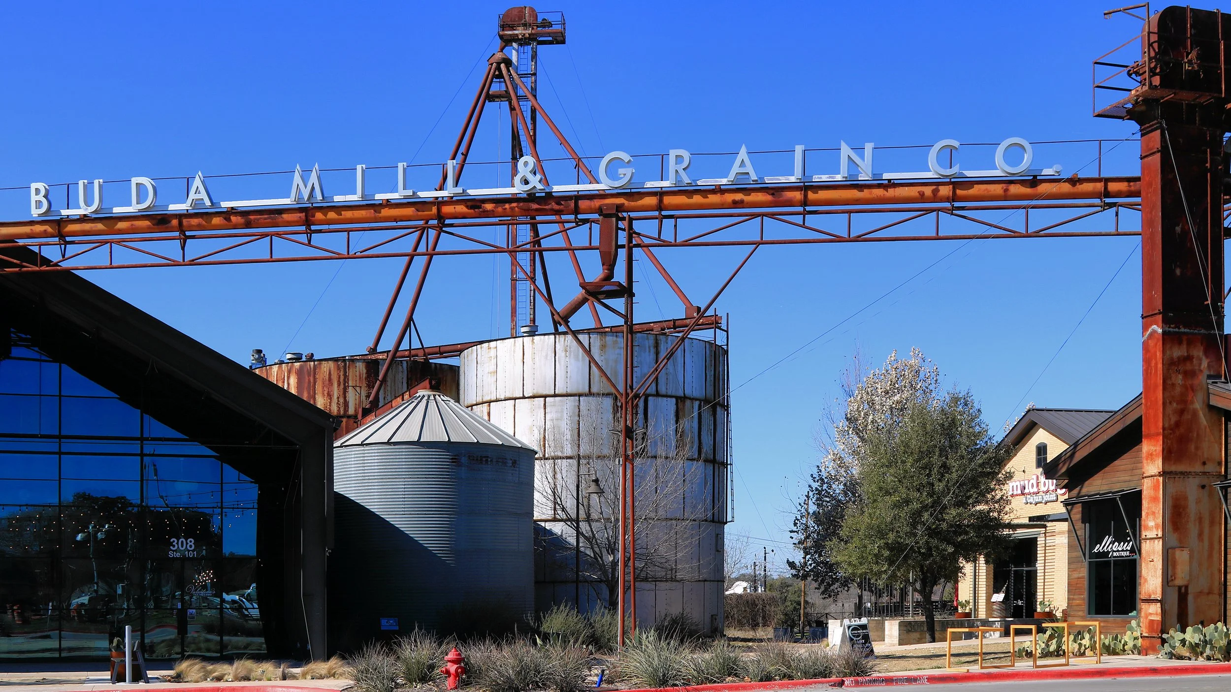 Rusty metal sign with white letters spelling 'BUDA MILL & GRAIN CO.' on an industrial building against a blue sky.