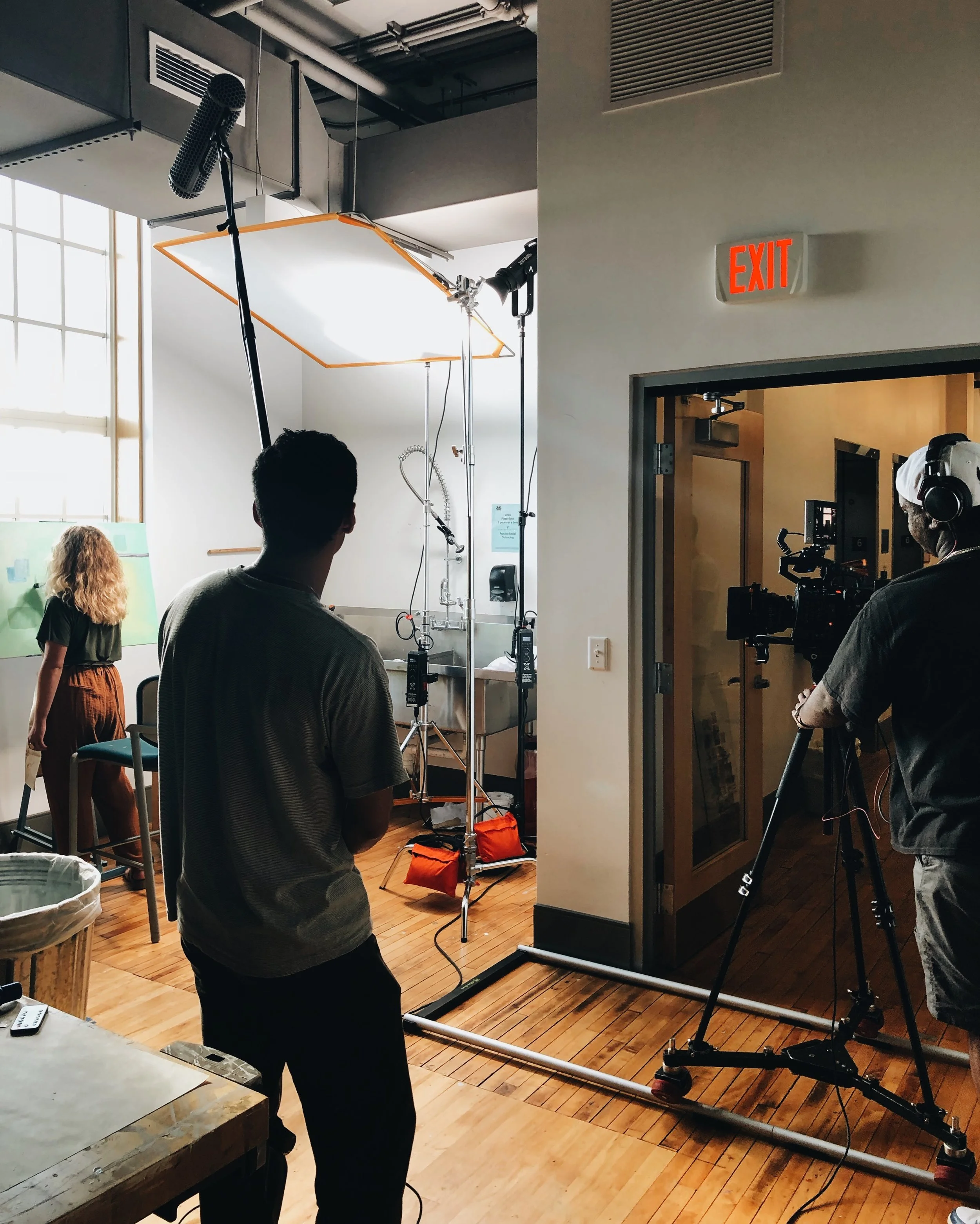 Behind the scenes of a video shoot in a bright studio space with crew members, camera equipment, lighting, and a woman with curly hair in the background.