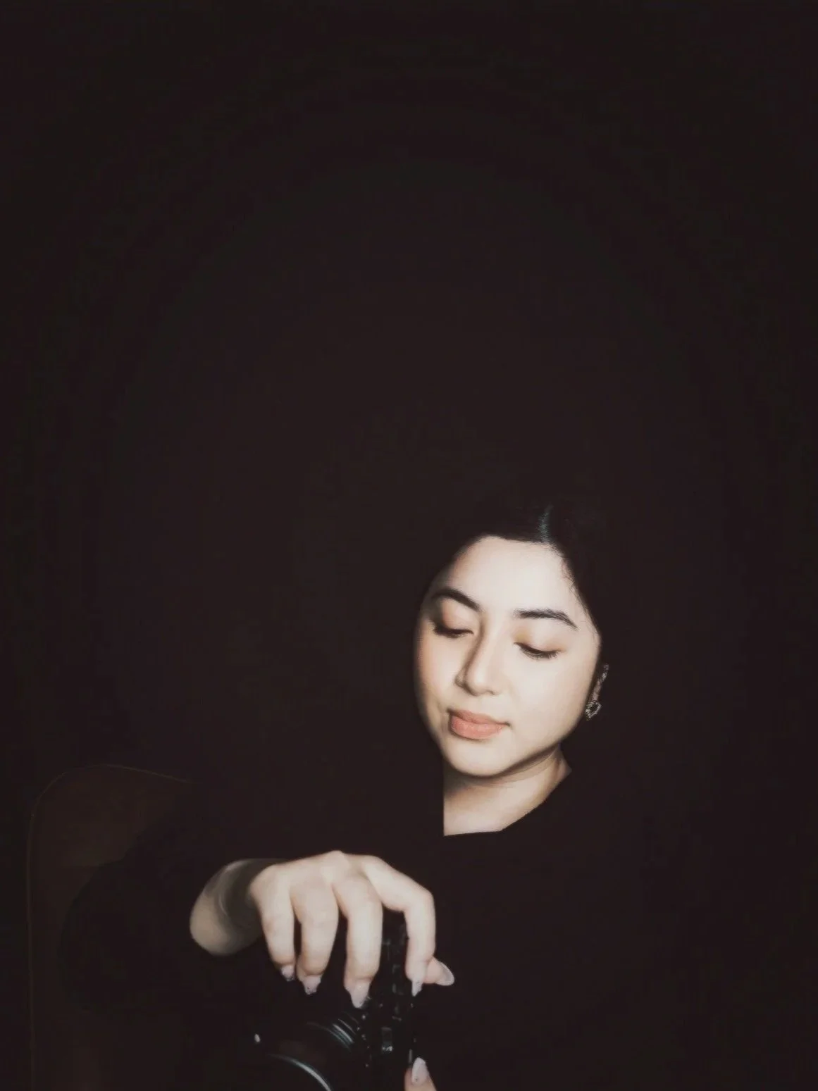 A woman with dark hair and earrings is taking a selfie with a smartphone, with her eyes closed against a dark background.