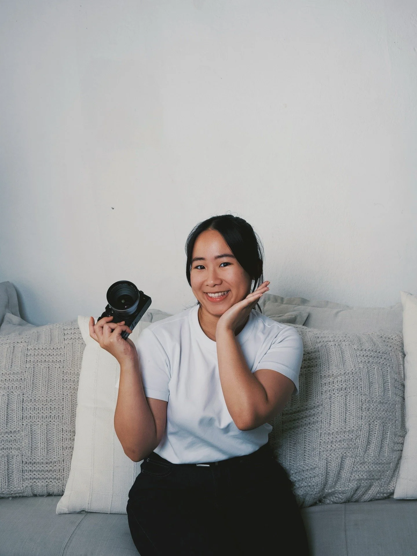 A smiling woman sitting on a sofa, holding a camera in her right hand and resting her left hand on her cheek.