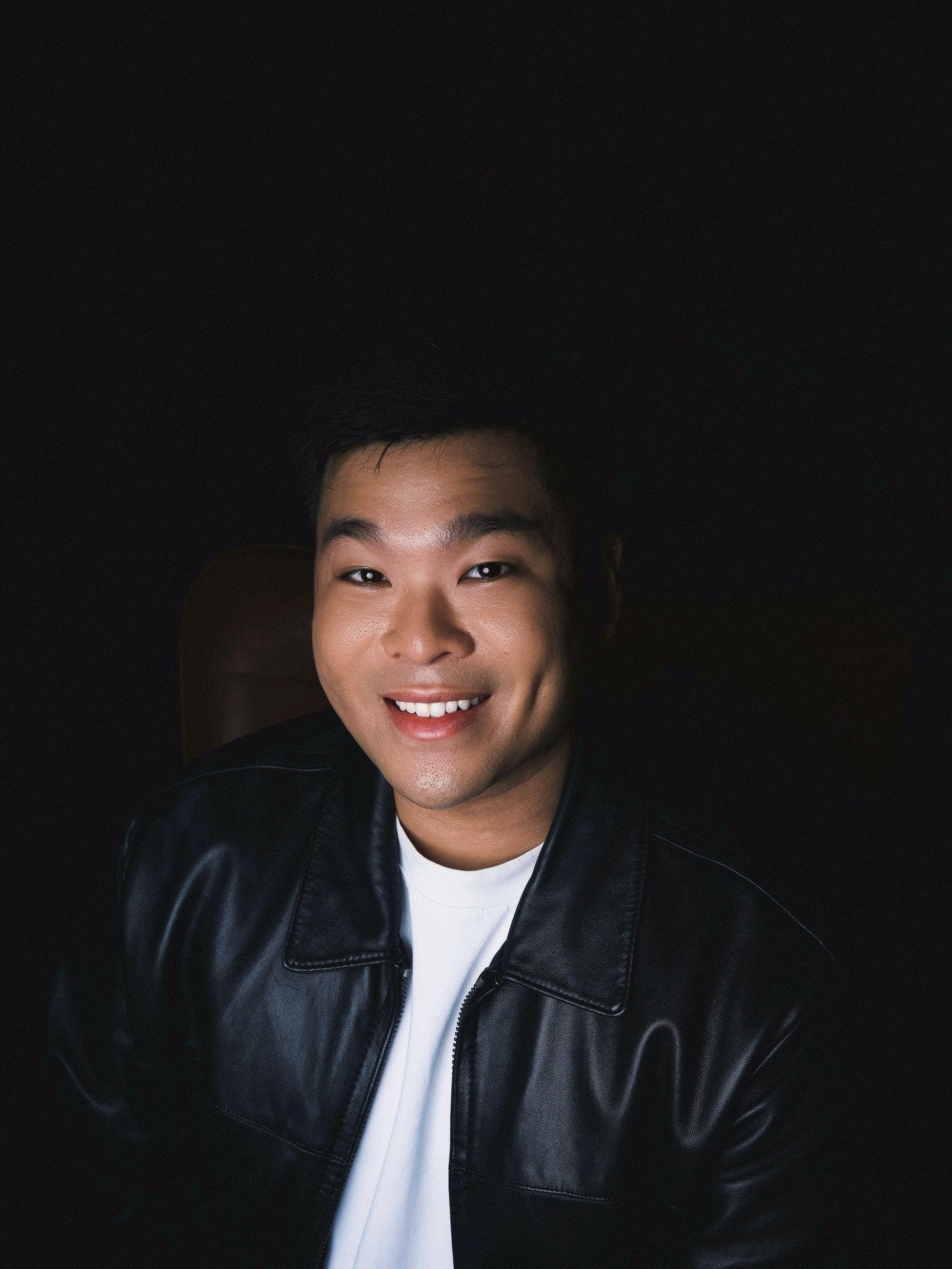 Close-up portrait of a smiling young man wearing a black leather jacket and white shirt, dark background.
