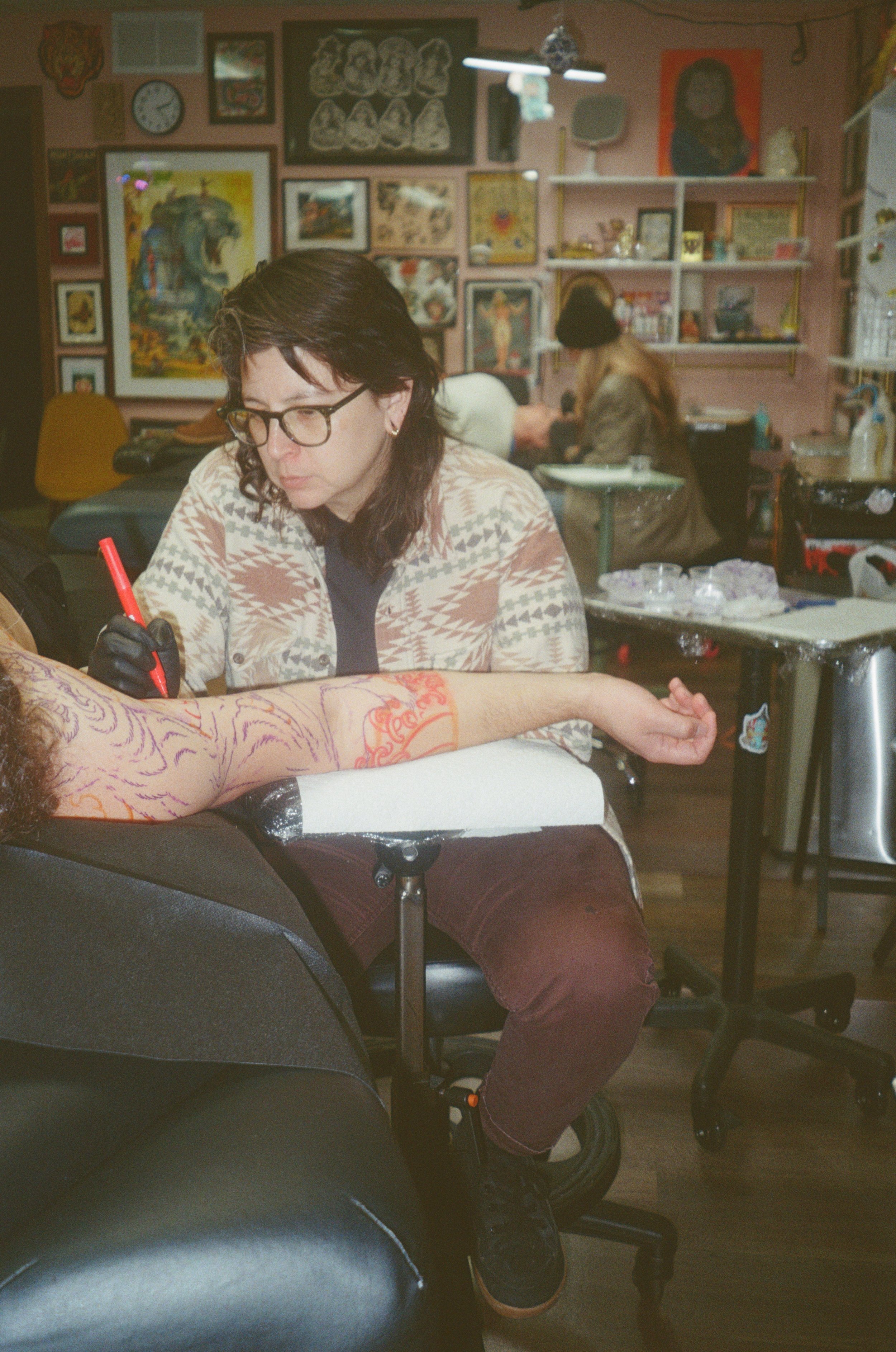 A woman with glasses and long dark hair standing in front of a bookshelf filled with books and art supplies. She is wearing a black T-shirt with a dragon tattoo design on the front, silver jewelry, and has colorful tattoos on her arms.