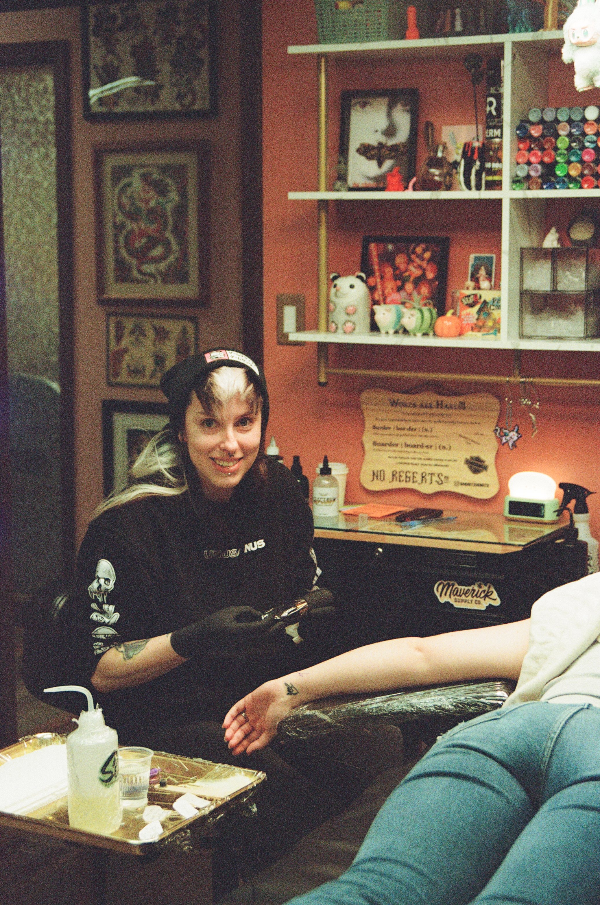 Tattoo artist with short, blonde and brown hair tattoos a person's skin using a blue tattoo gun in a tattoo studio.