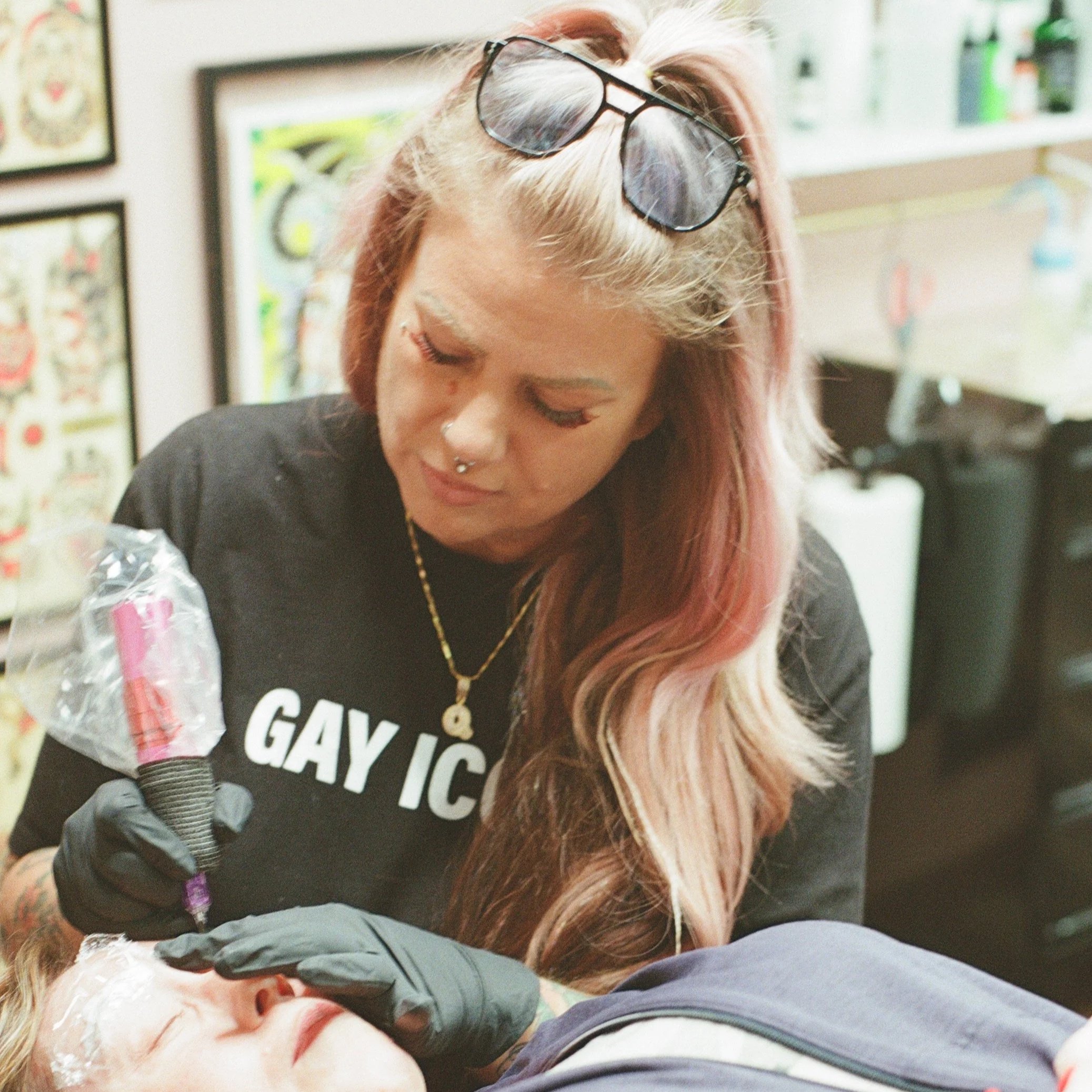 A smiling woman with blonde hair, tattoos, and multiple rings, wearing a black jacket with butterfly embroidery, sitting against a wooden background.