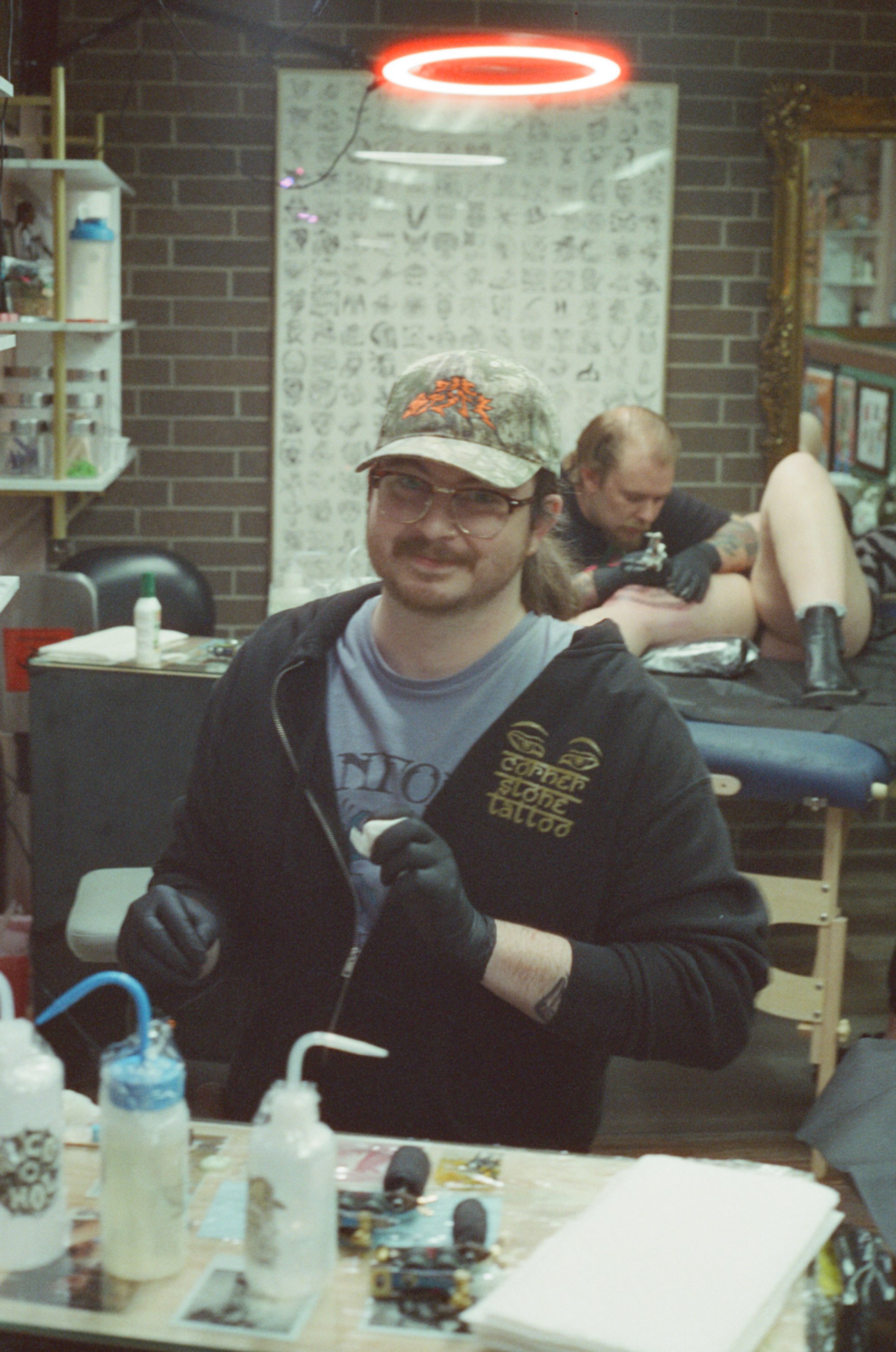 A tattooed man wearing glasses, a beige cowboy hat, and a pink shirt is getting a tattoo from a tattoo artist in a tattoo studio decorated with artwork and tattoo supplies.