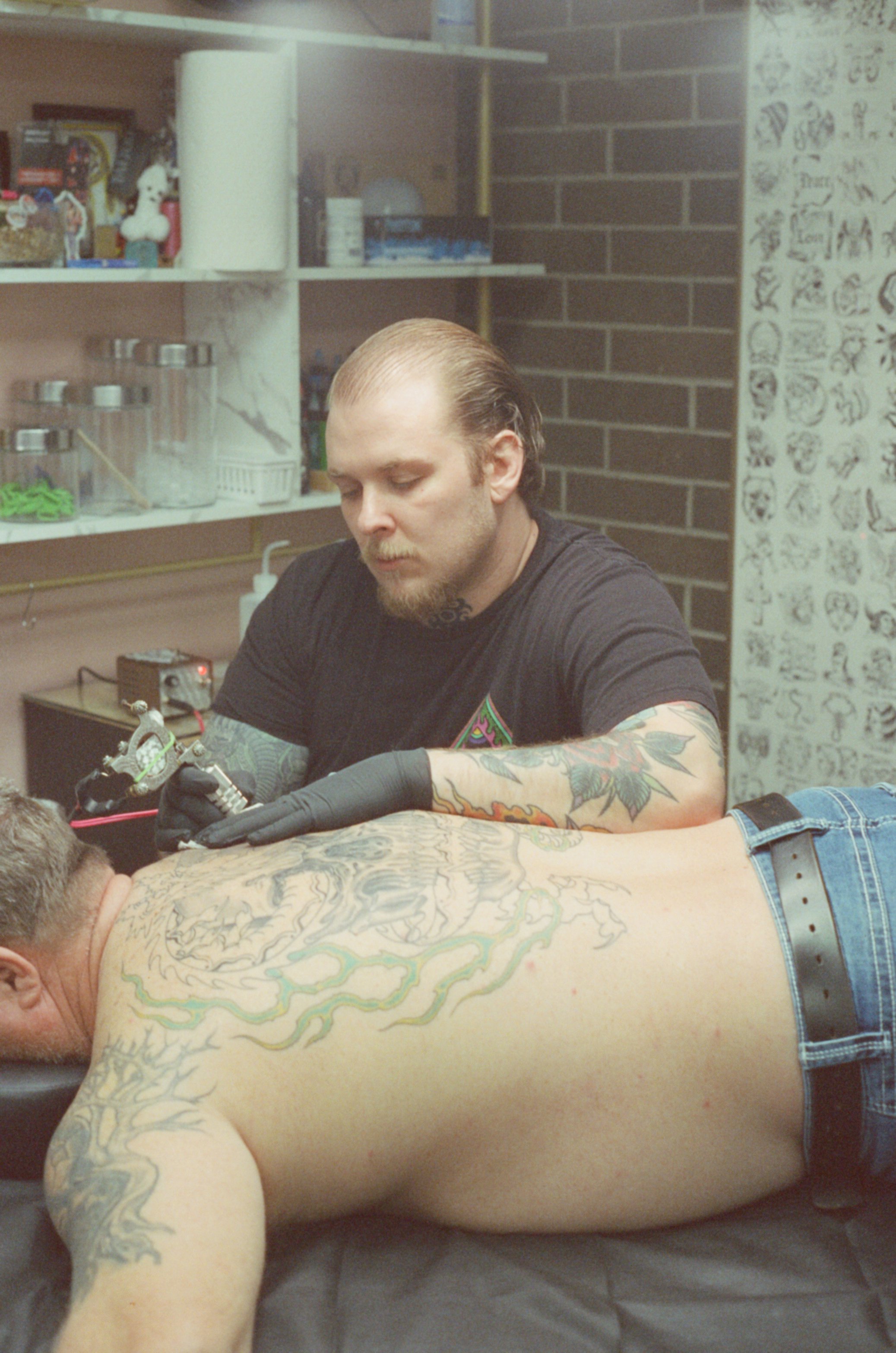 Tattoo artist tattooing a client's arm in a tattoo studio with brick wall background.