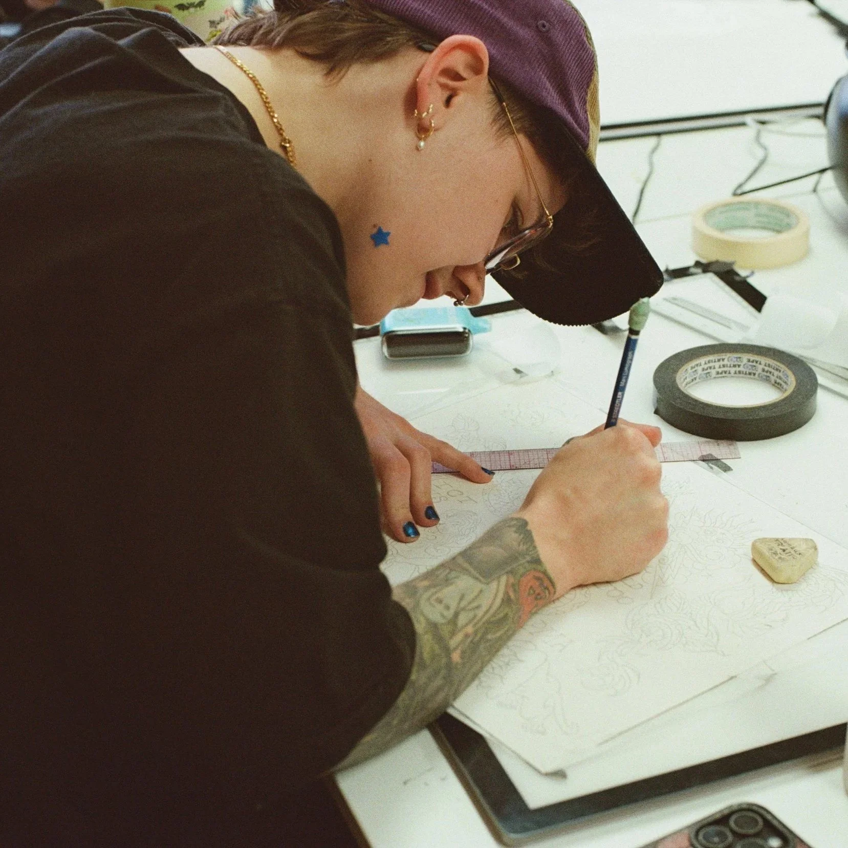 A person with curly hair, glasses, and tattoos is drawing a tattoo design on paper, sitting at a table with art supplies. There are bookshelves filled with books in the background.
