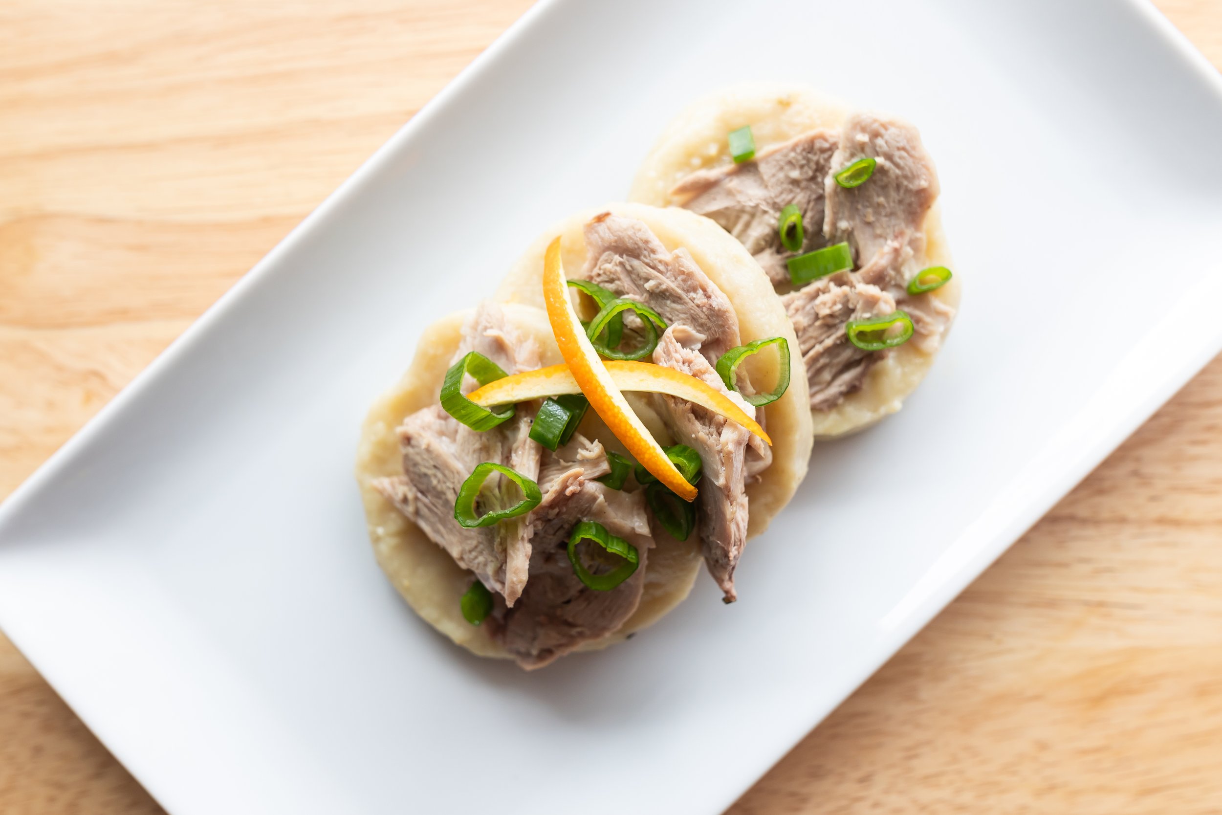 Two bao buns topped with pulled pork, sliced green onions, and orange zest on a white rectangular plate.