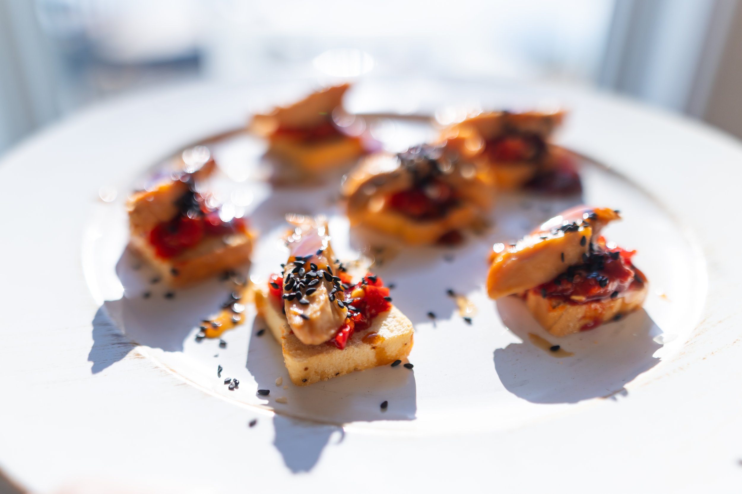 Gourmet appetizers on a plate with toasted bread, topped with red sauce and sesame seeds.