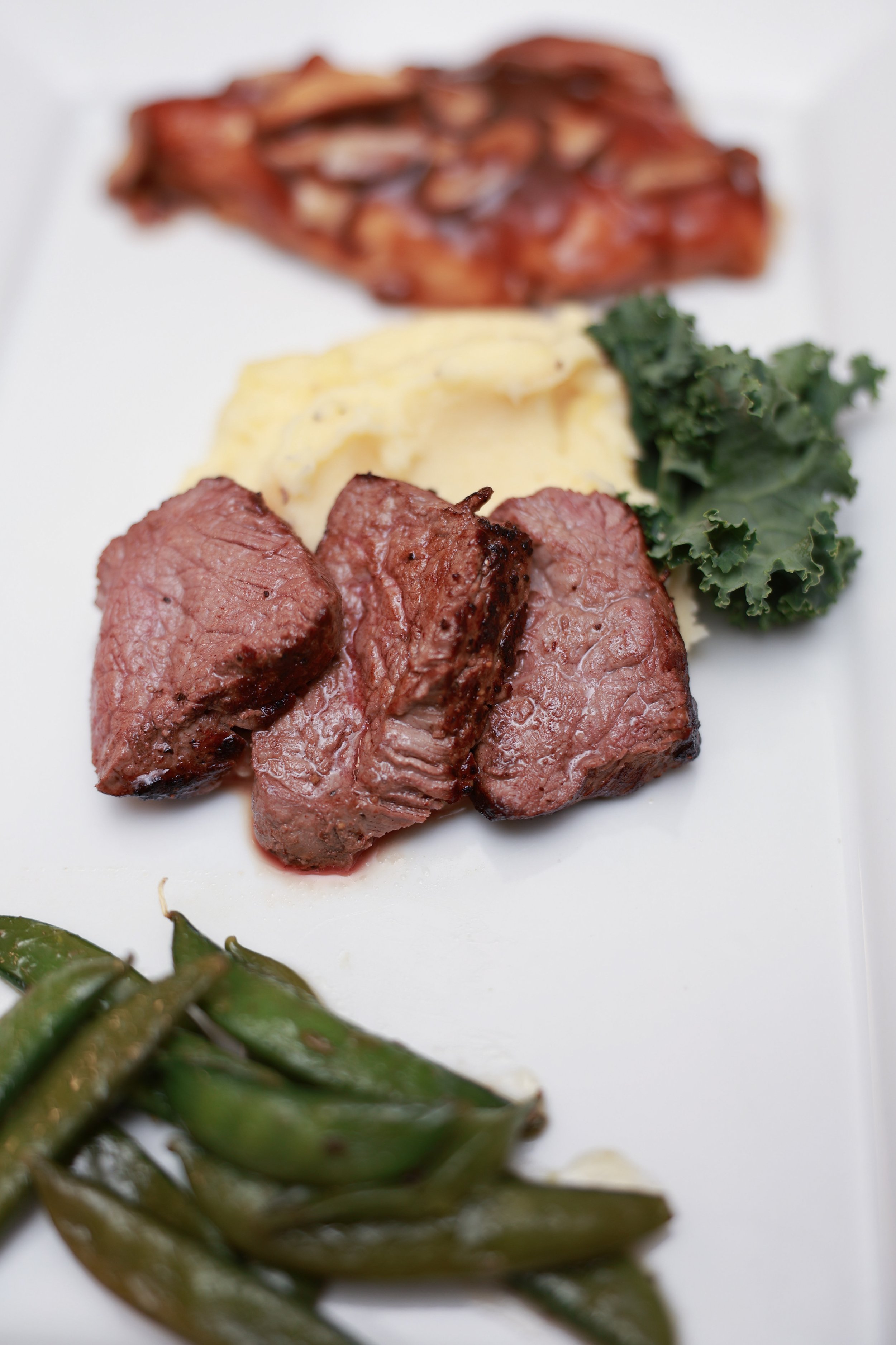 Slices of grilled beef with mashed potatoes, kale, glazed carrots, and sugar snap peas on a white plate.