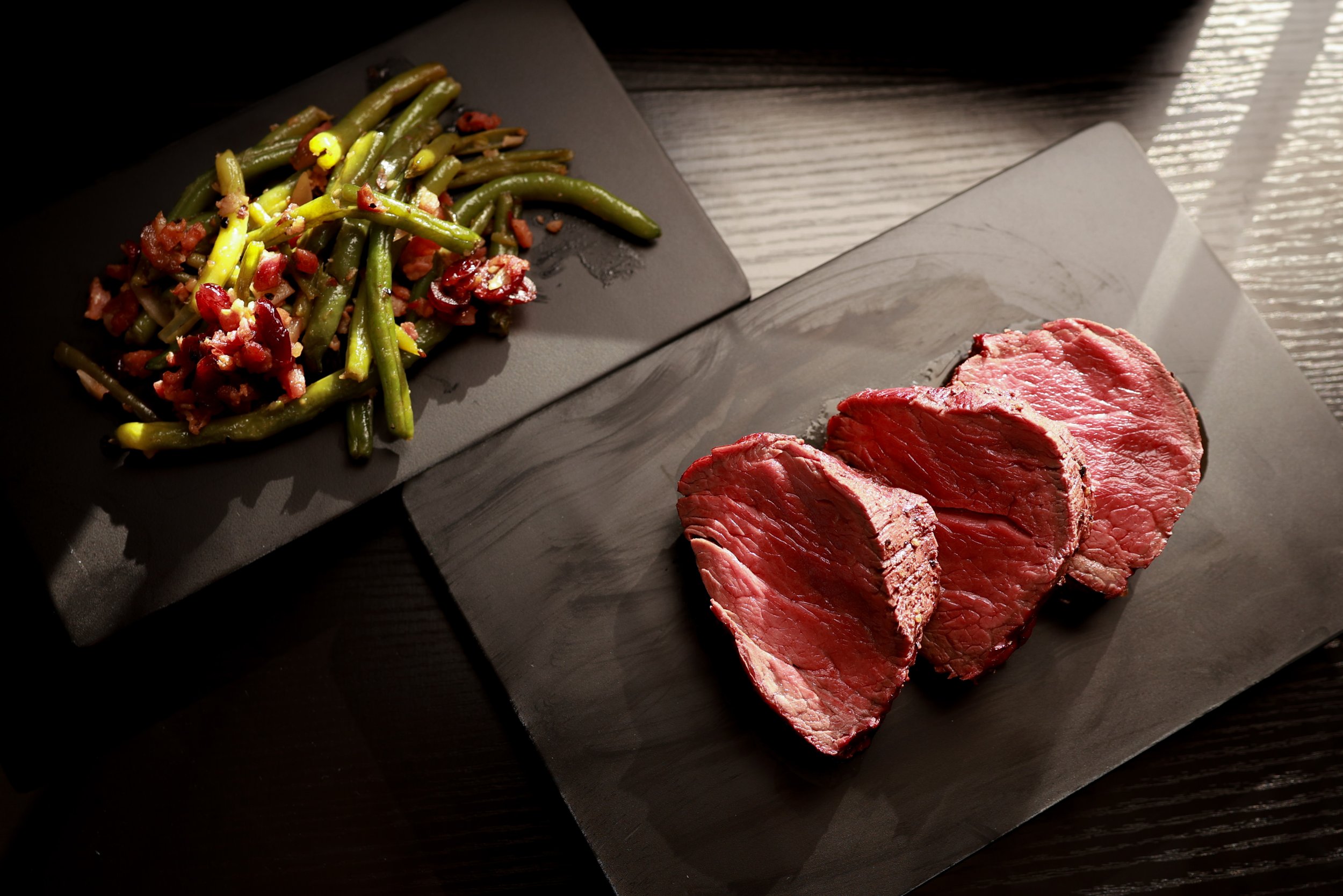 Sliced beef steak and green beans with bacon and red berries on black plates.