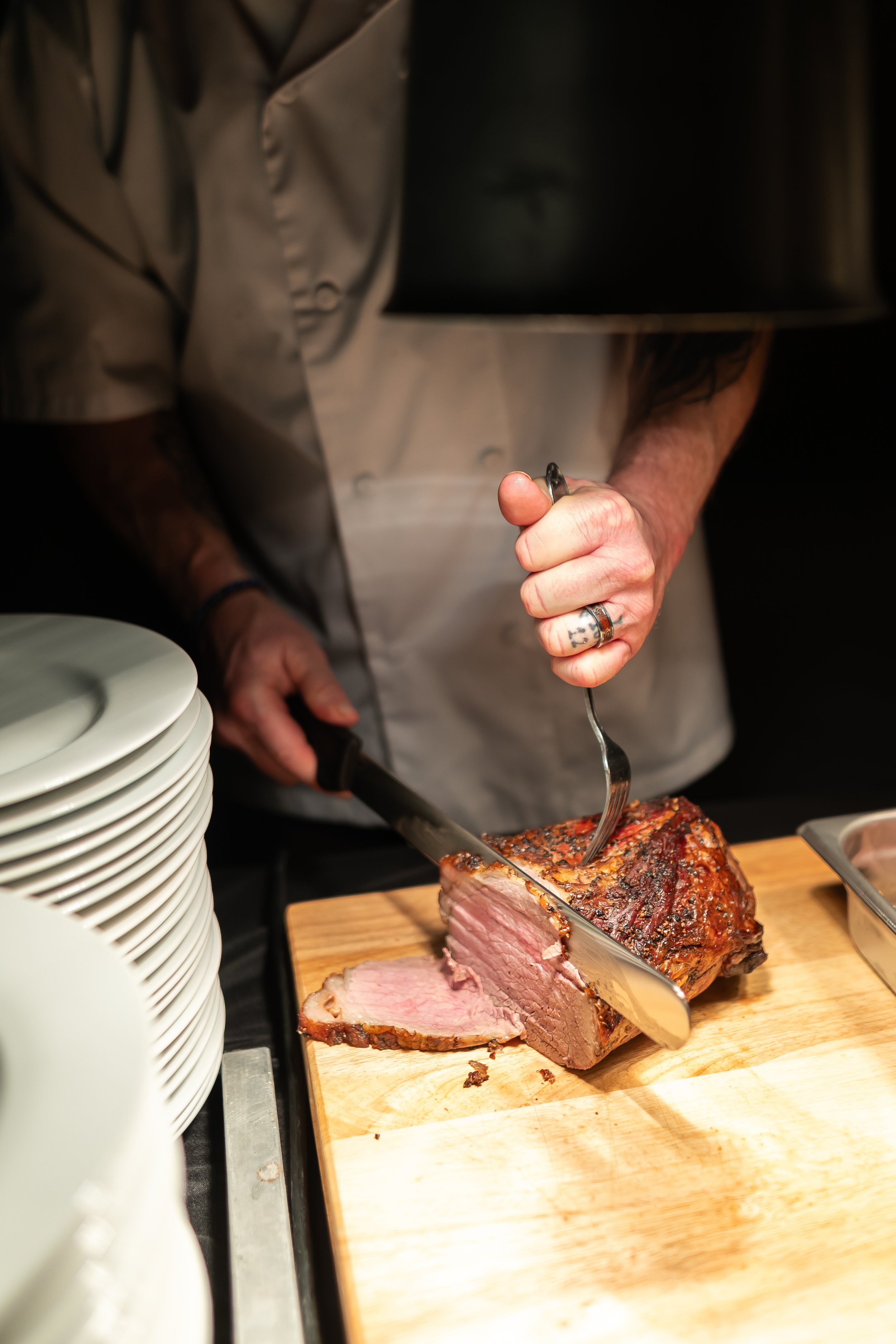 Chef carving a piece of roasted meat on a wooden board with a knife and fork.